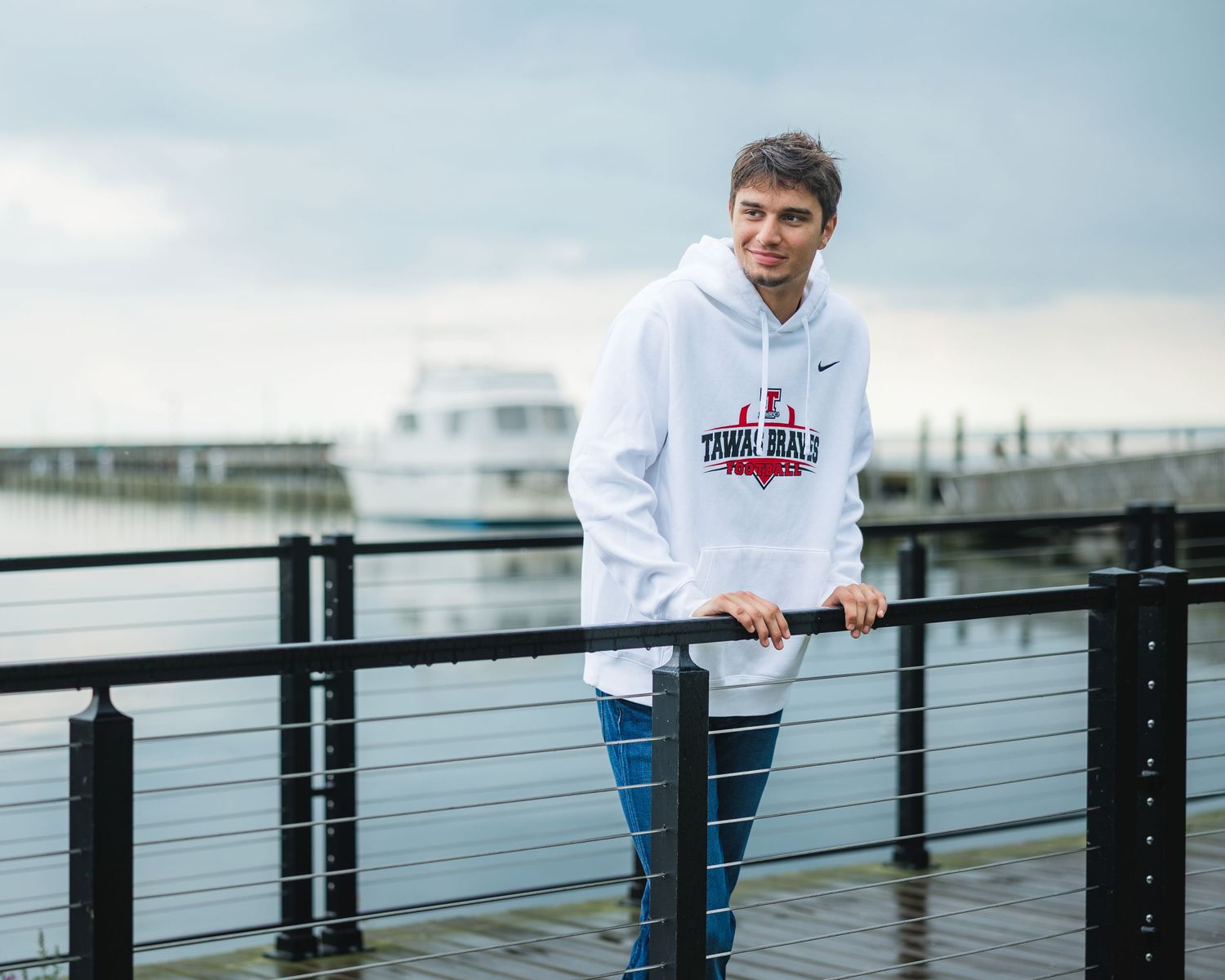Young man in white Nike hoodie posing on waterfront dock with marina and overcast sky in background.