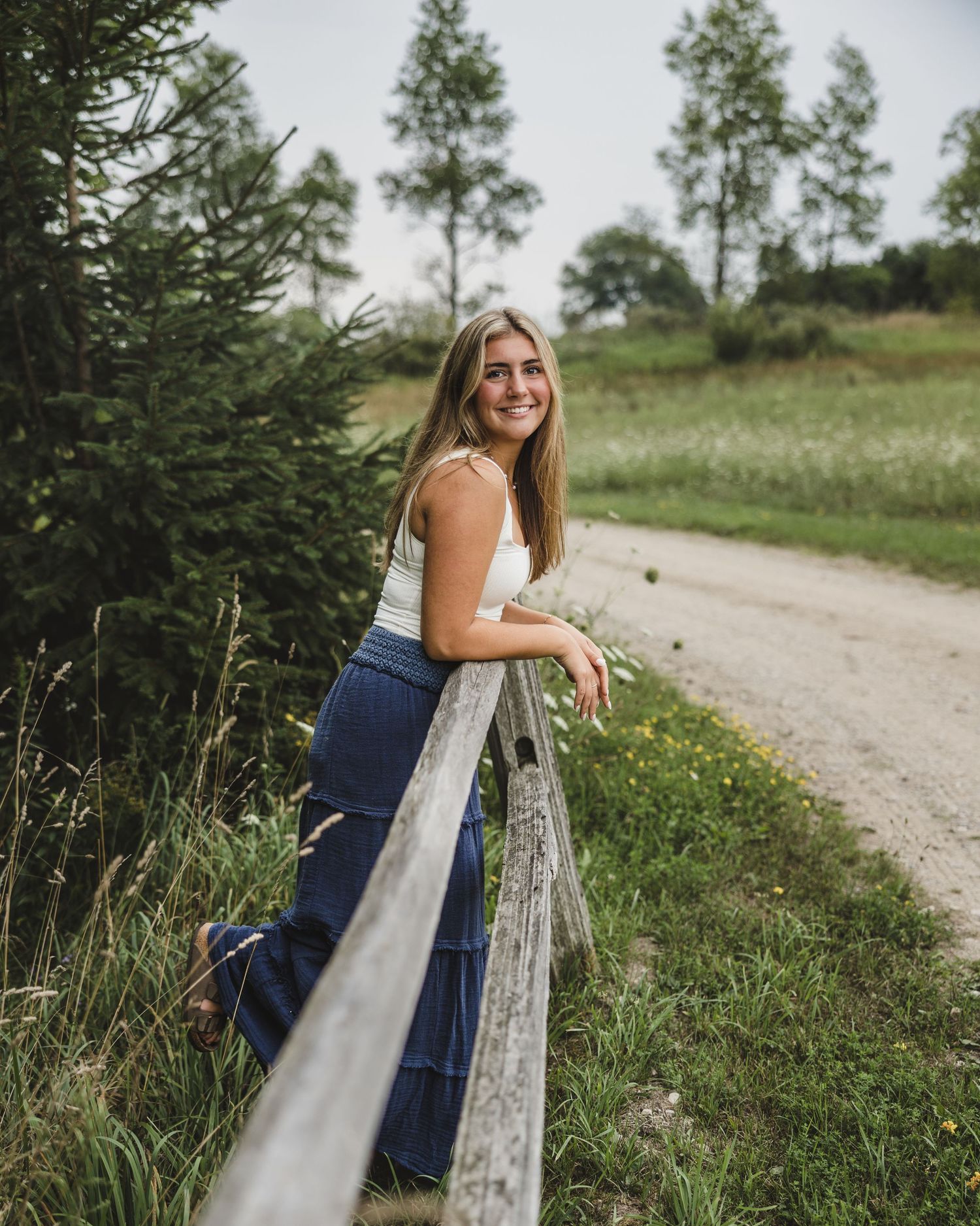 Young woman in white top and blue flared pants smiling while leaning on a rustic fence along a country road.