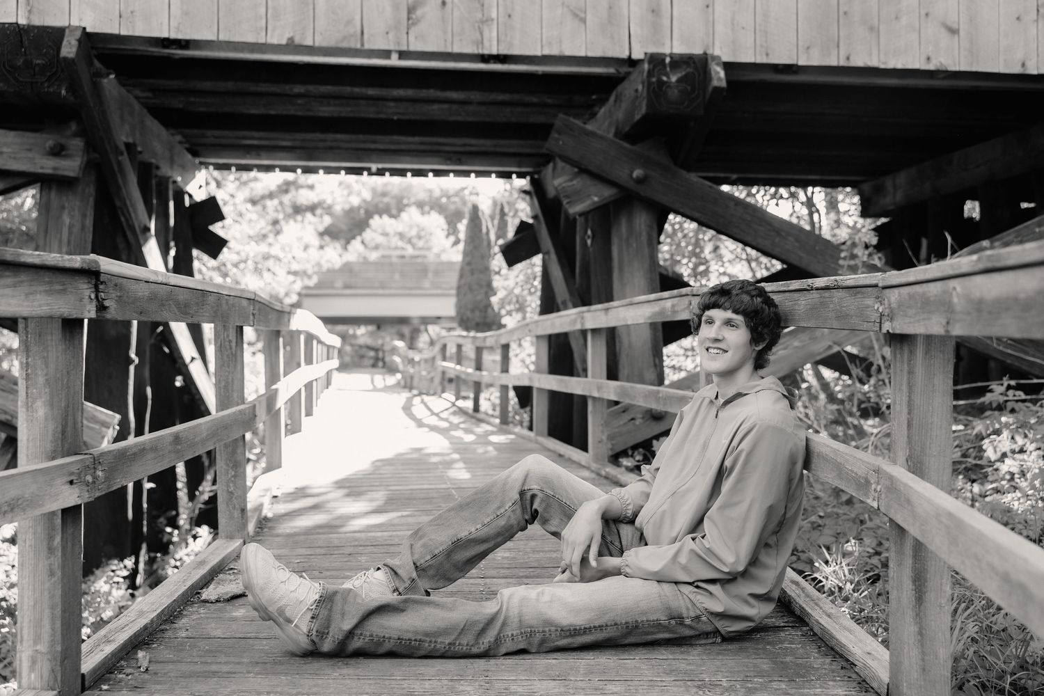 Young person relaxing on a wooden covered bridge, smiling casually in a black and white outdoor photo.