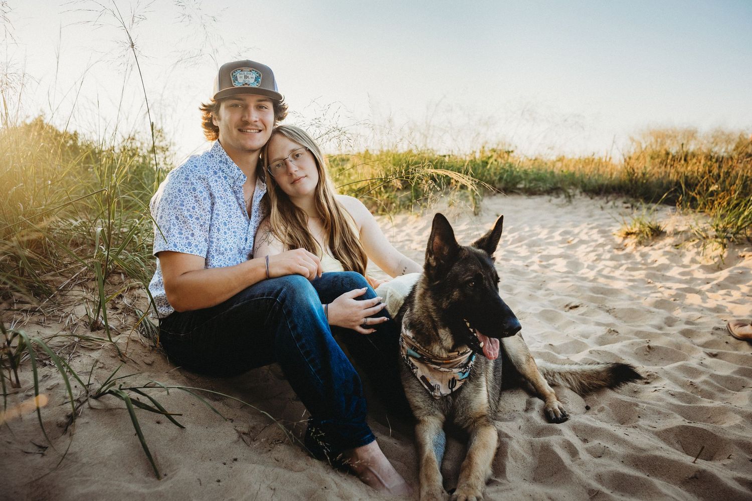 A couple sits with their German Shepherd dog on a sandy beach, smiling together in warm golden light.