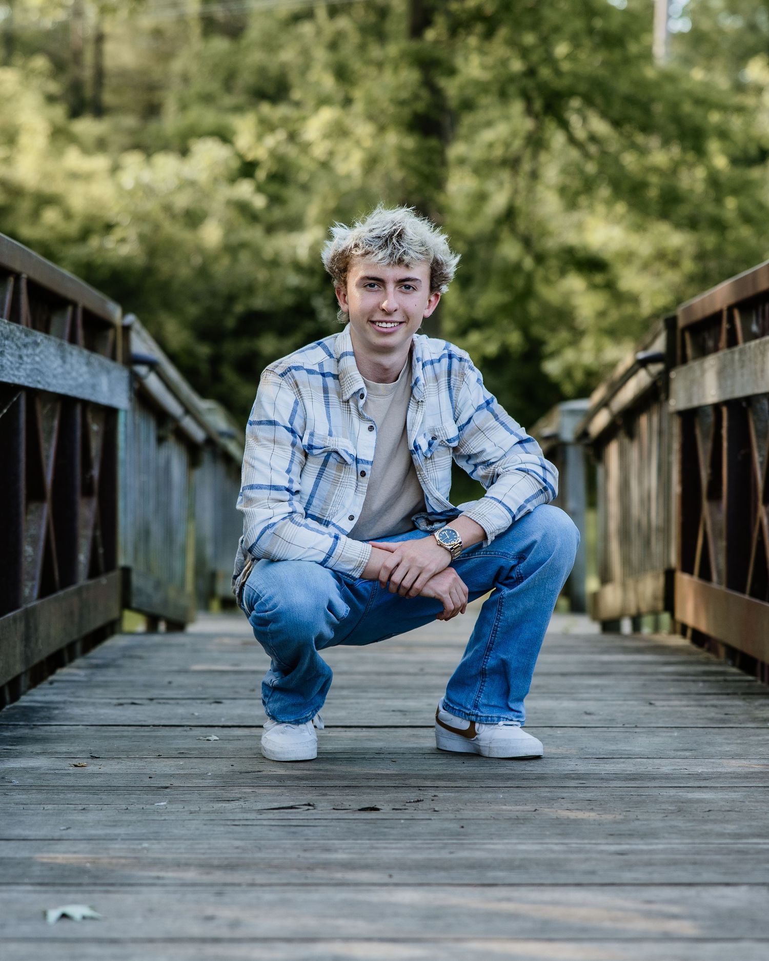 Smiling young man in plaid shirt squatting on wooden bridge surrounded by green trees.