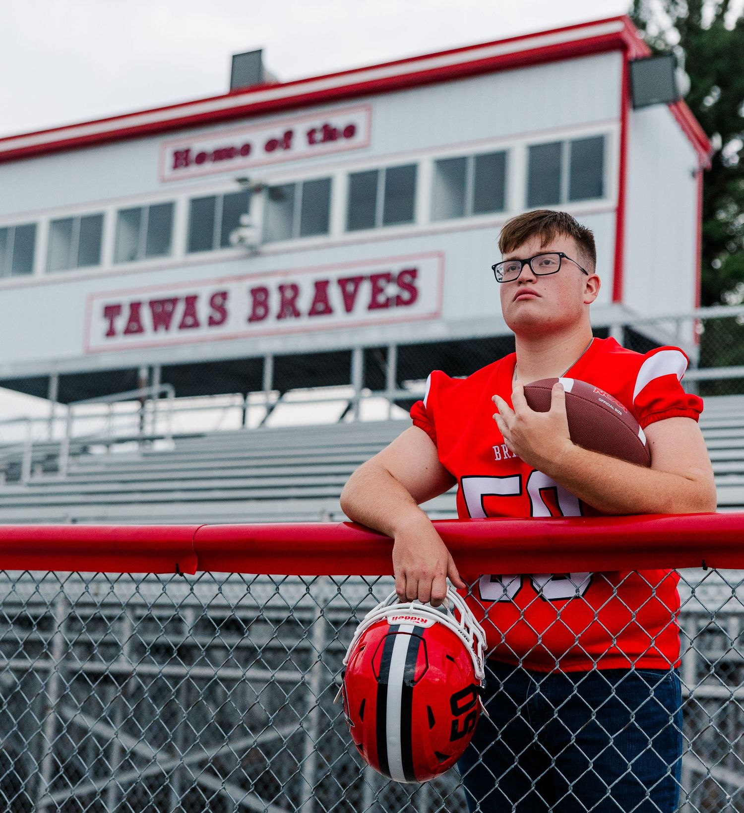 Young football player in red Tawas Braves uniform holding helmet and football at team stadium.