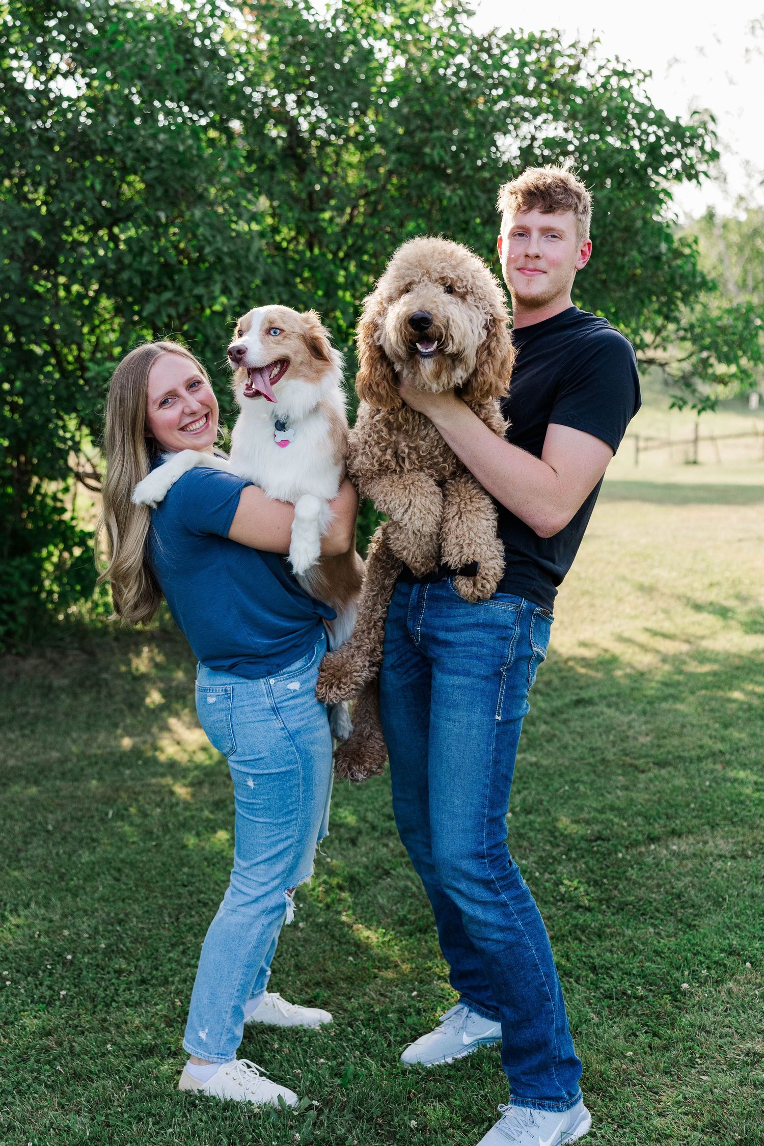 A smiling couple holds two dogs, an Australian Shepherd and a Goldendoodle, outdoors on green grass.