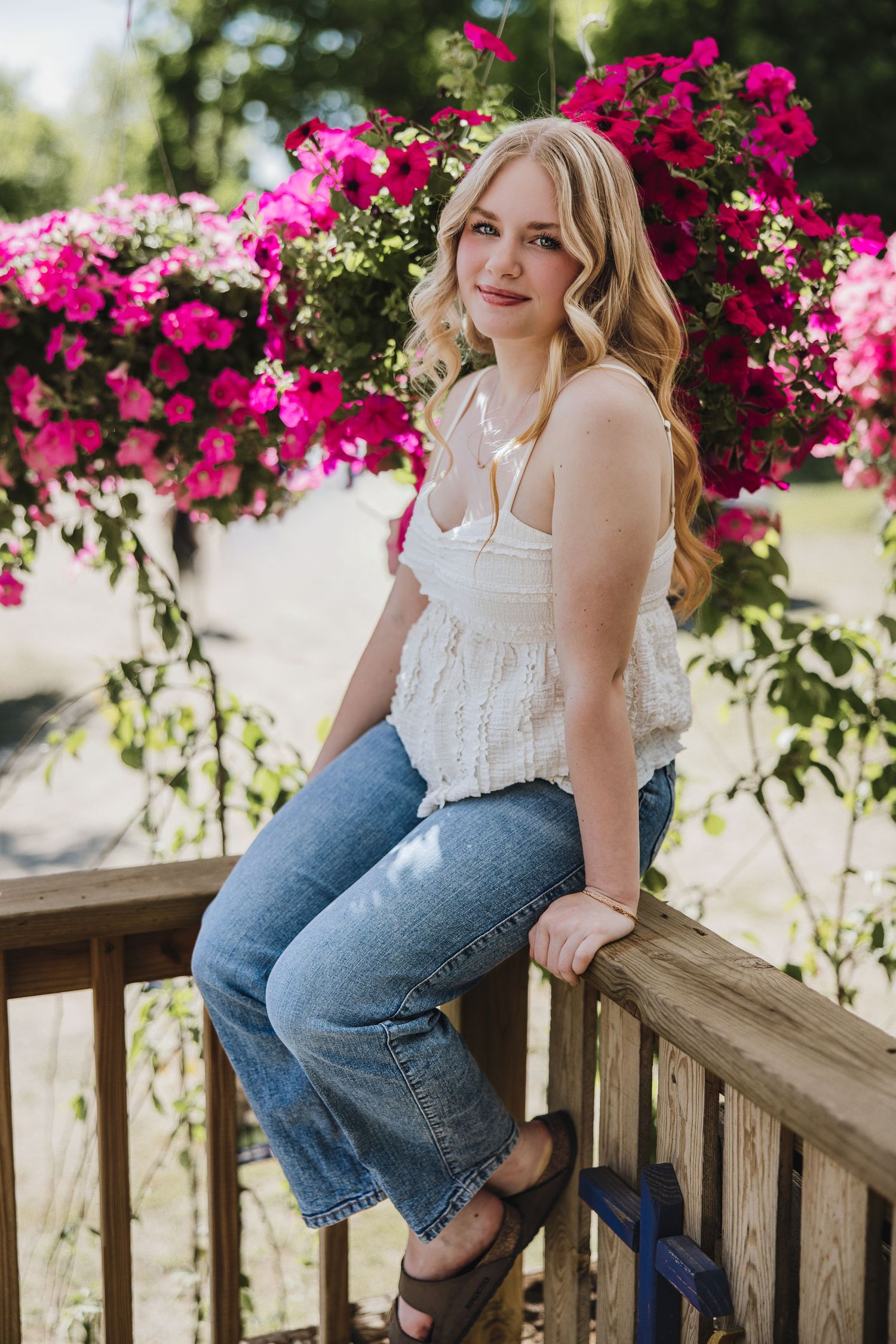 Young blonde woman in white top and jeans sitting on wooden deck railing with vibrant pink flowers behind her.