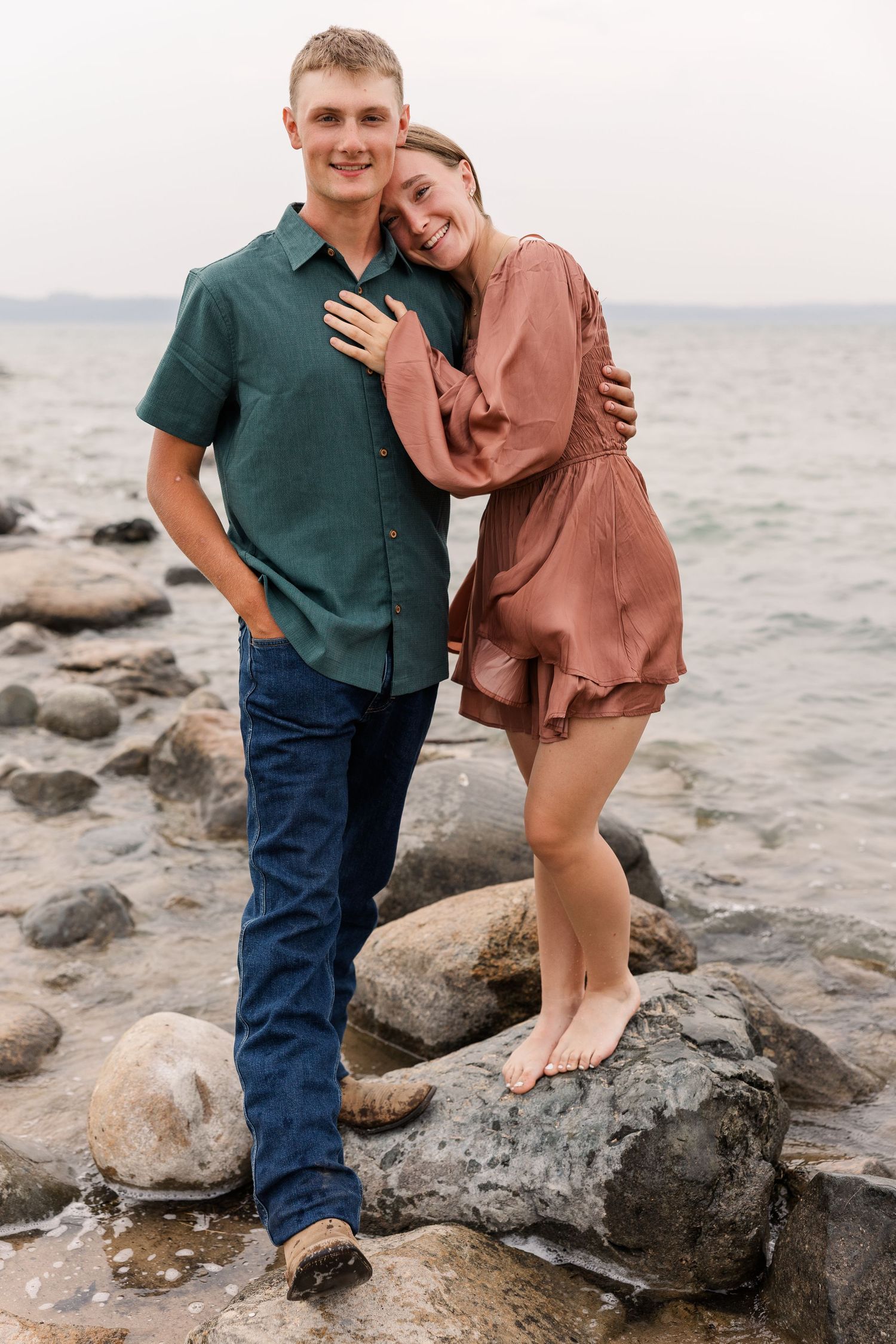 Couple embracing on rocky shoreline, woman in mauve dress leaning on man in green shirt and jeans, smiling by the sea.