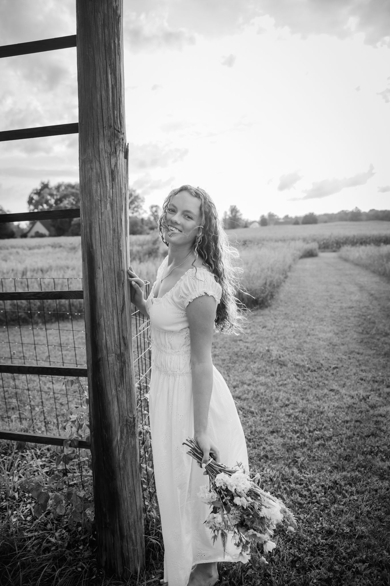 Young woman in white dress holding bouquet stands by rustic fence post in black and white farm field photo.