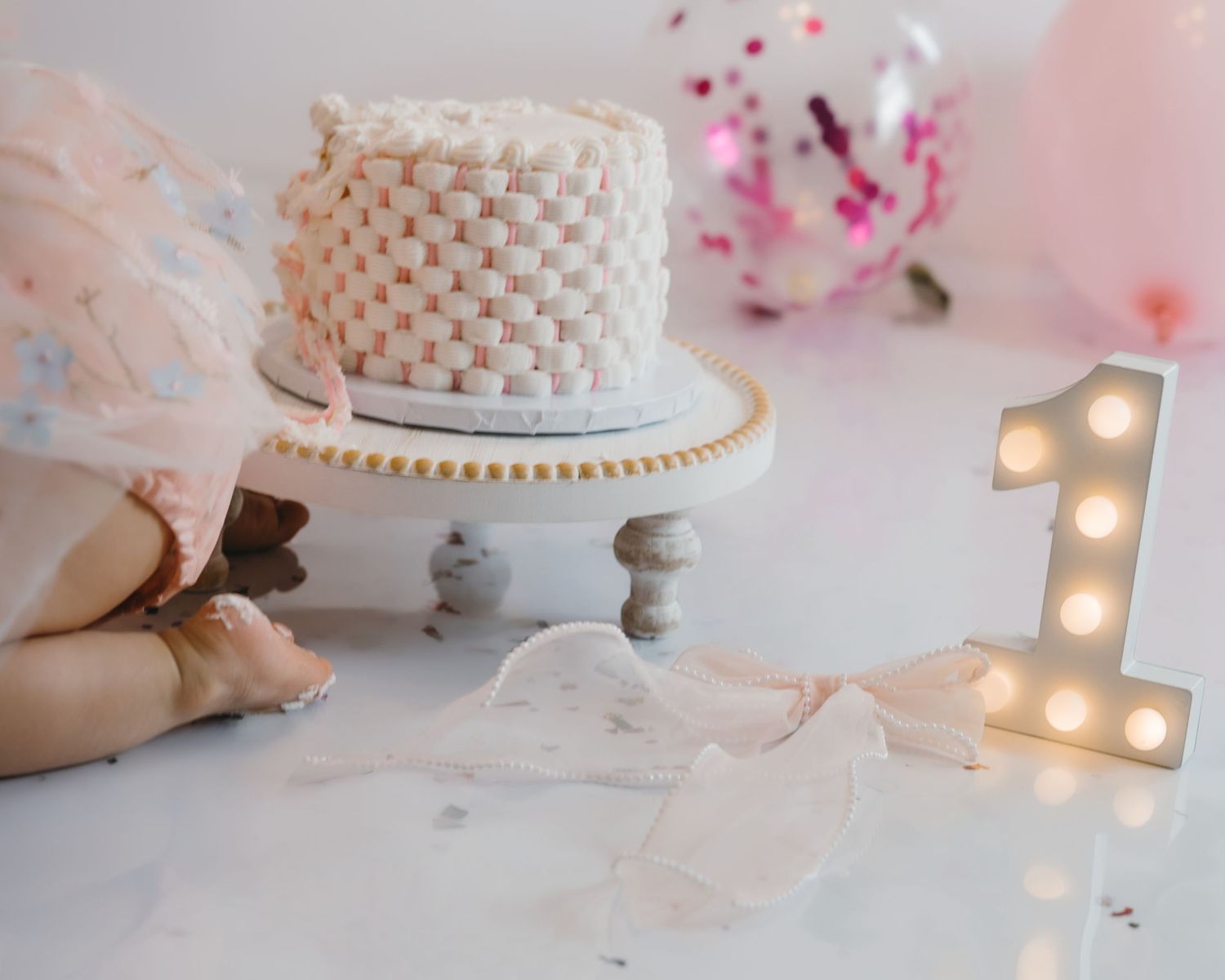 Baby in pink tutu dress reaches toward elegant white basket-weave first birthday smash cake.