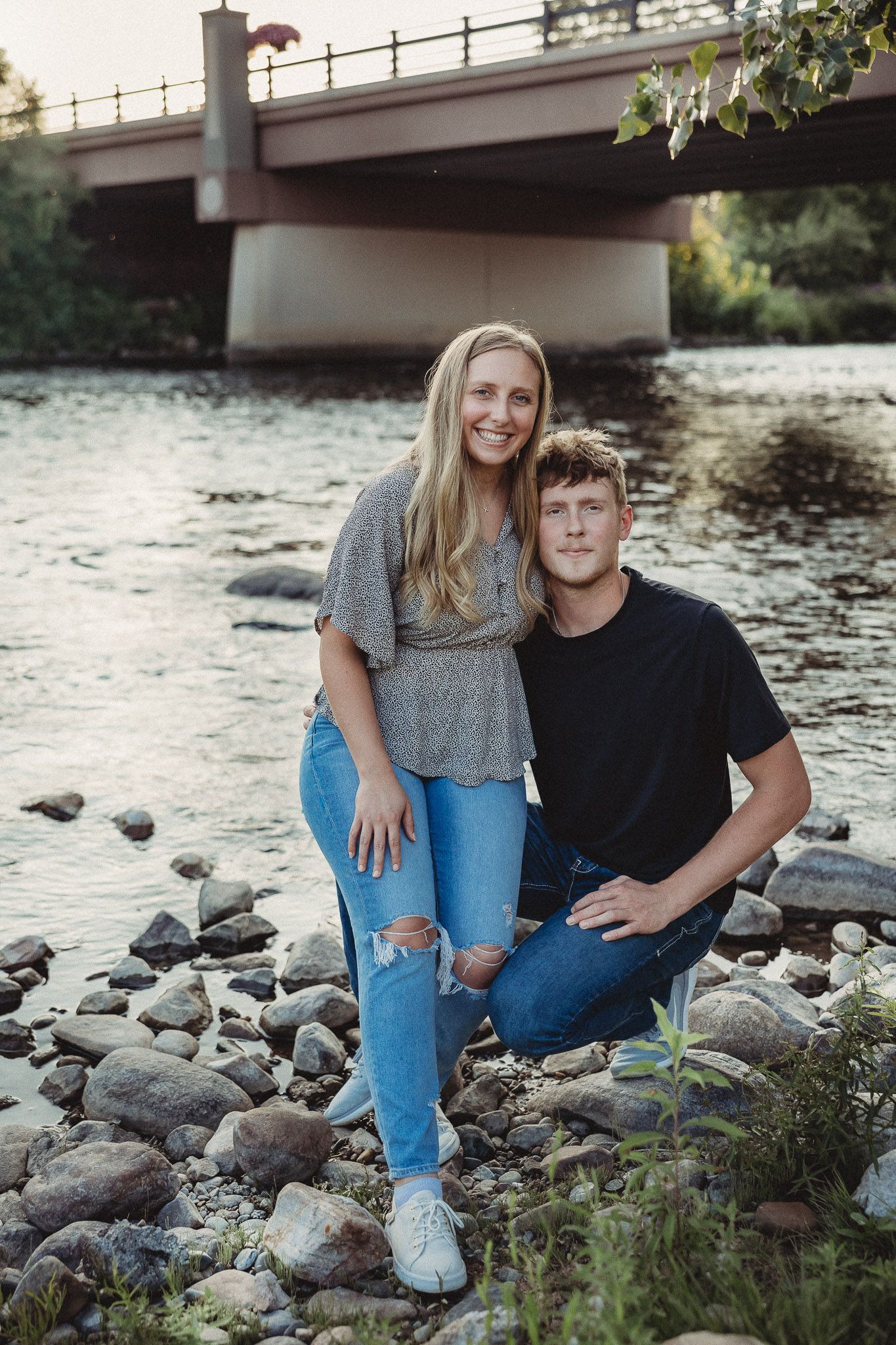 Smiling couple posing by a rocky riverbank with a bridge in the background.