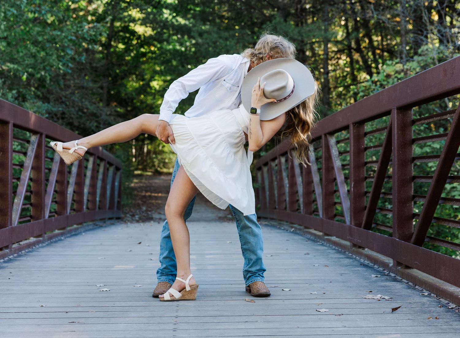Couple sharing romantic dip kiss on wooden bridge surrounded by lush green forest trees.