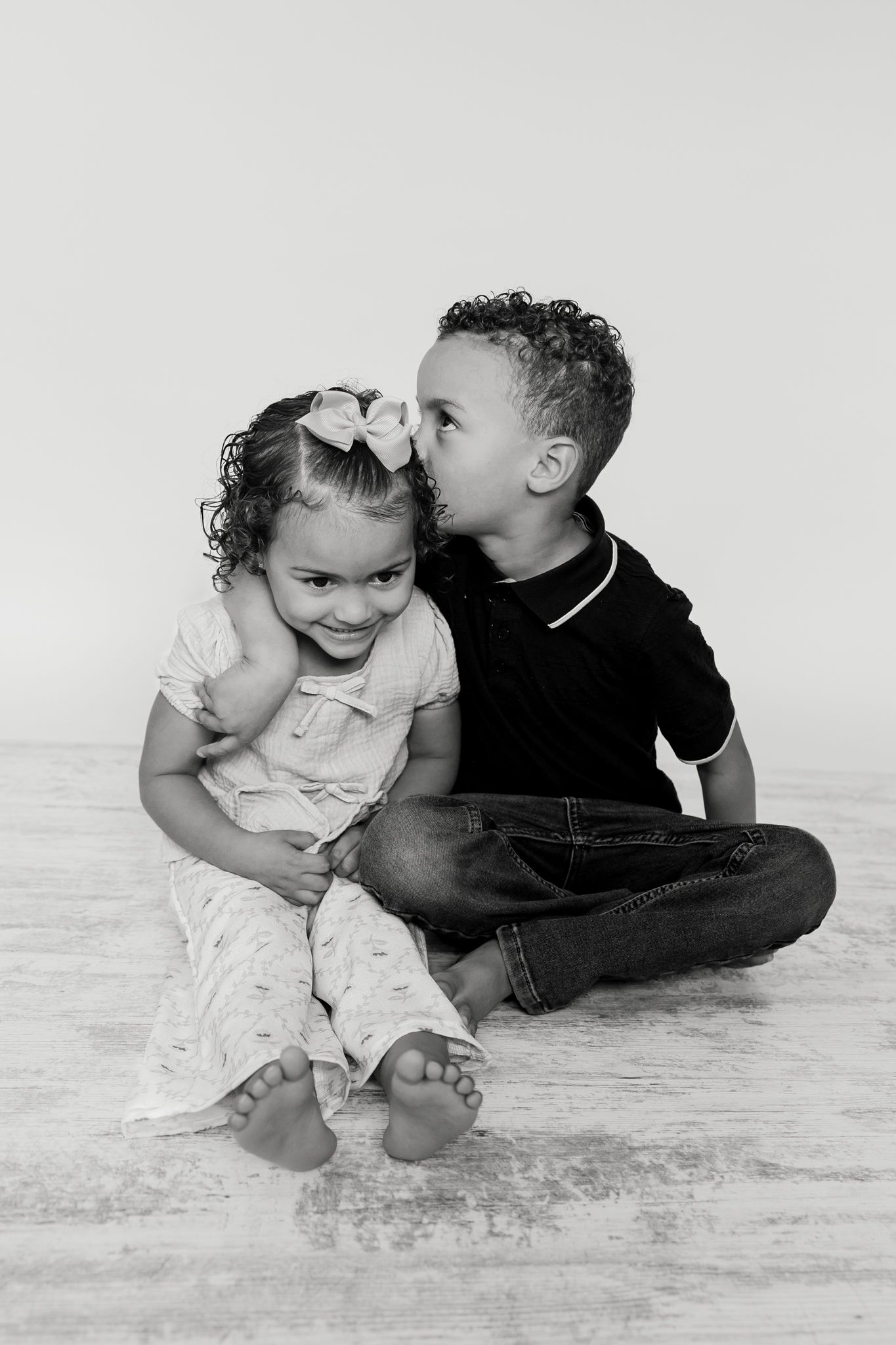 Black and white photo of two young siblings sitting together, older boy kissing younger girl on the head.