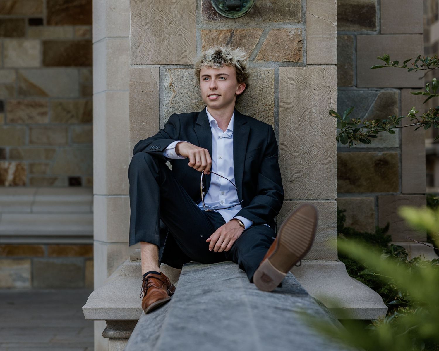 Young man in dark blazer and dress pants sitting on stone ledge outdoors near stone columns.