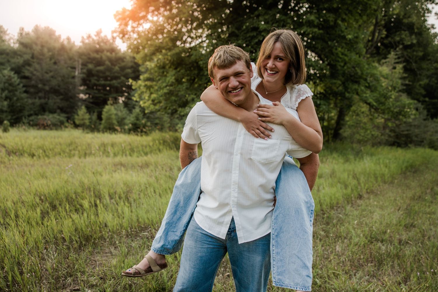 Couple laughing outdoors, man giving woman a piggyback ride in a green meadow with trees in the background.