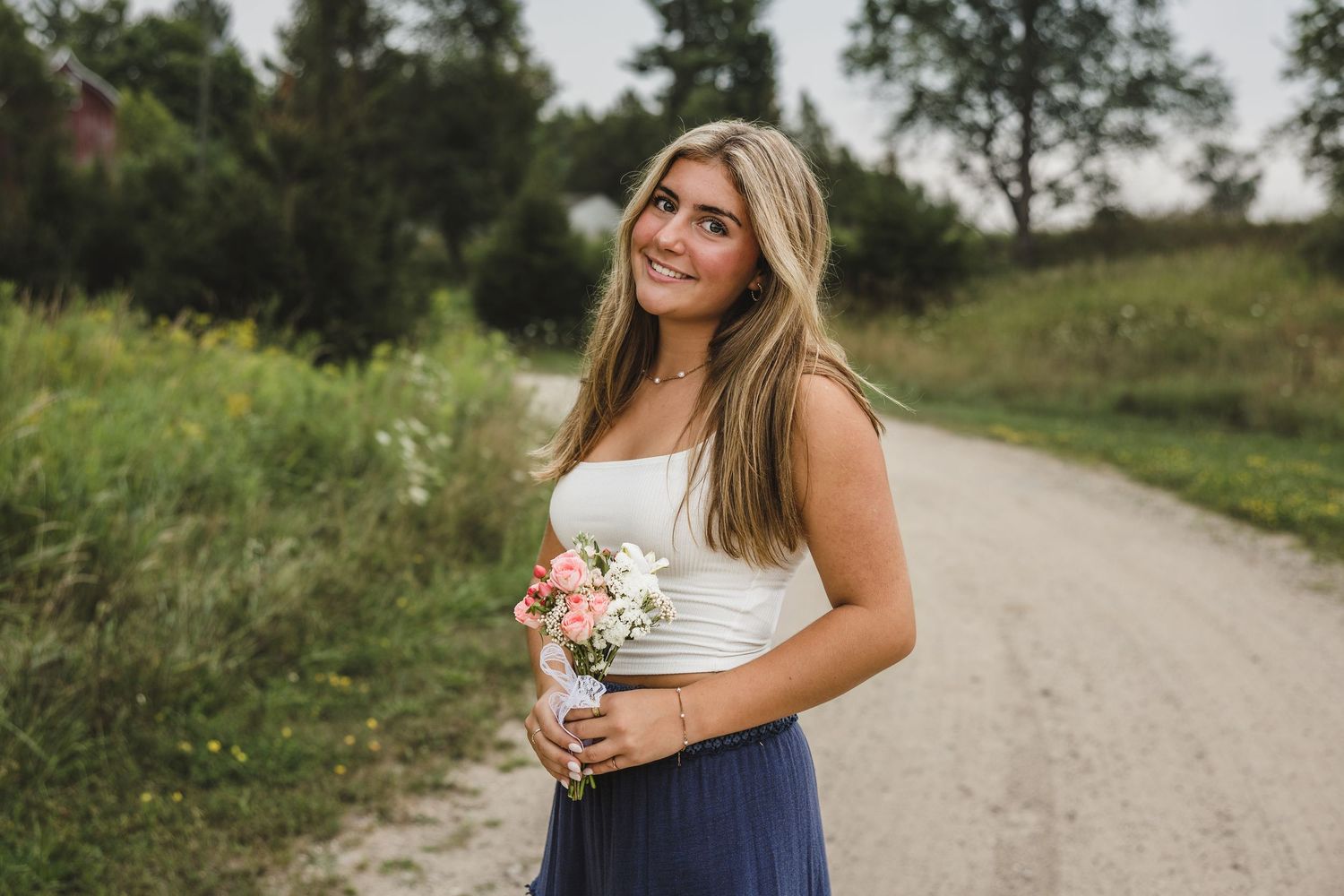 Young woman in white top and blue skirt holding a small floral bouquet on a scenic gravel path.