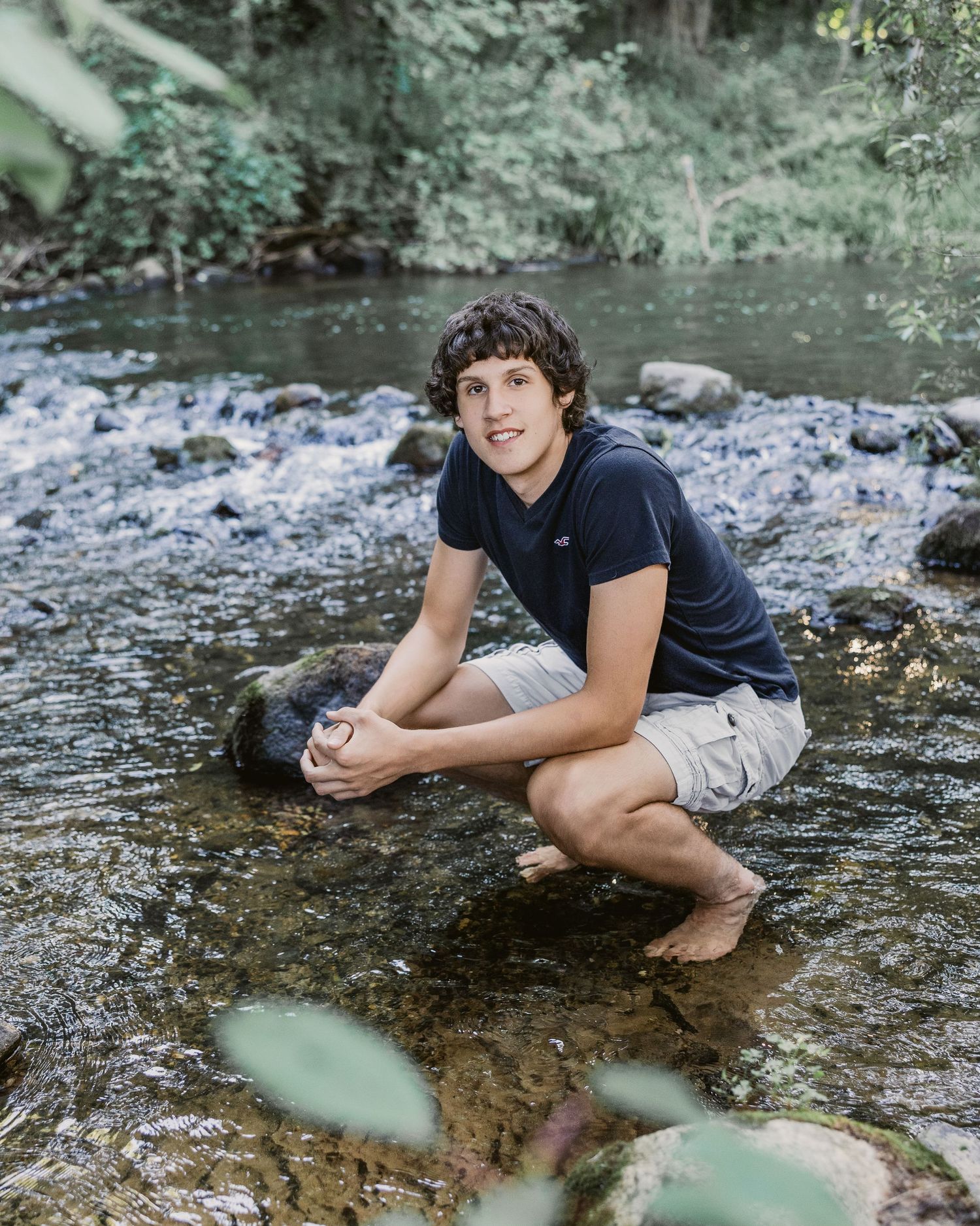 A teenage boy crouches barefoot in a rocky creek, wearing khaki shorts and a navy shirt, surrounded by lush green trees.