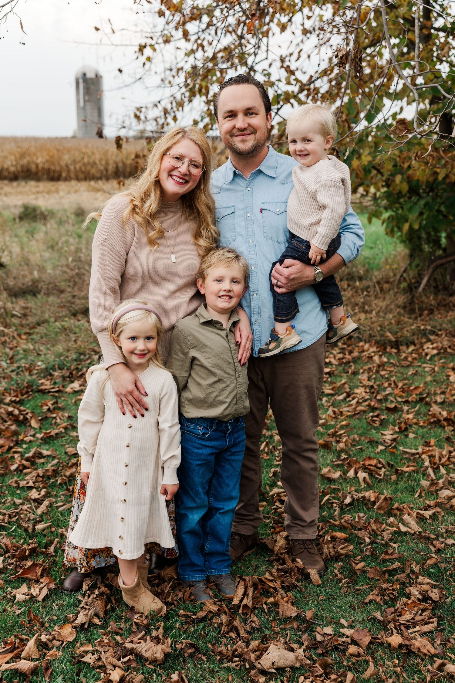 A happy family of five poses outdoors among fall leaves near a farm field.