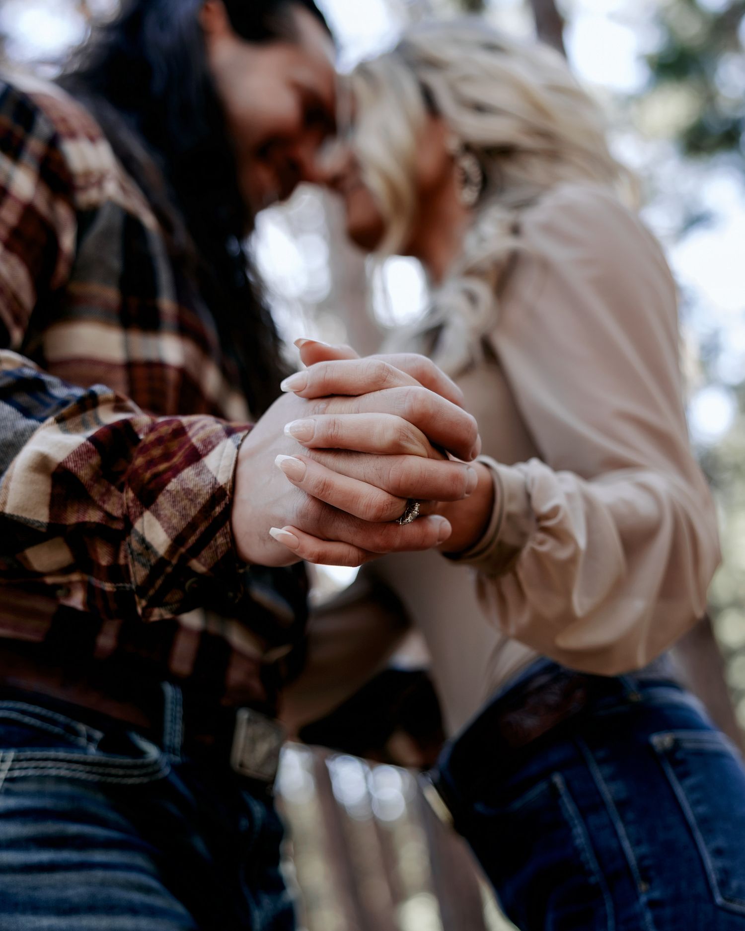 Couple holding hands tenderly in a forest, wearing flannel and cream blouse, with a ring visible on their intertwined fingers.