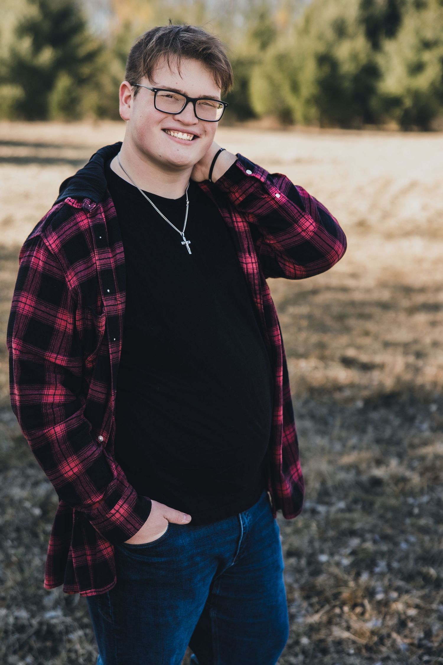 Smiling young man in red plaid shirt, black hoodie, jeans, and glasses wearing a cross necklace outdoors.