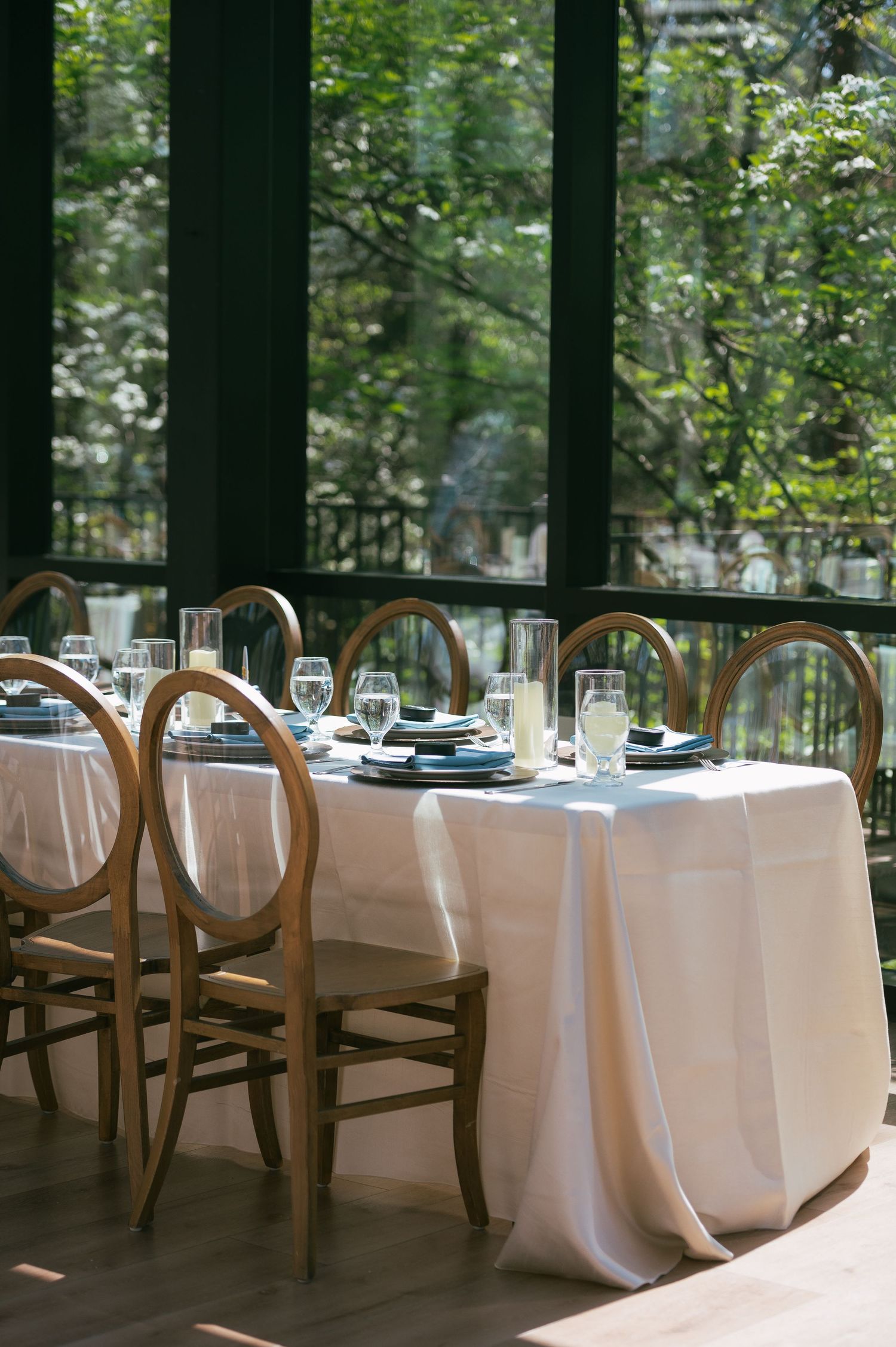Elegant dining table with white linen, blue plates, and wood circle-back chairs beside large windows overlooking lush greenery.