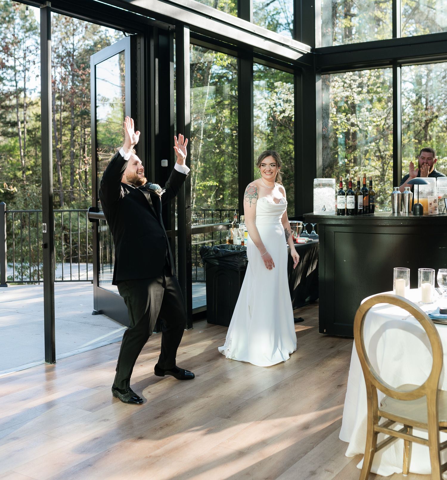 Groom raises hands in celebration as bride in white gown smiles in elegant glass-walled wedding venue.