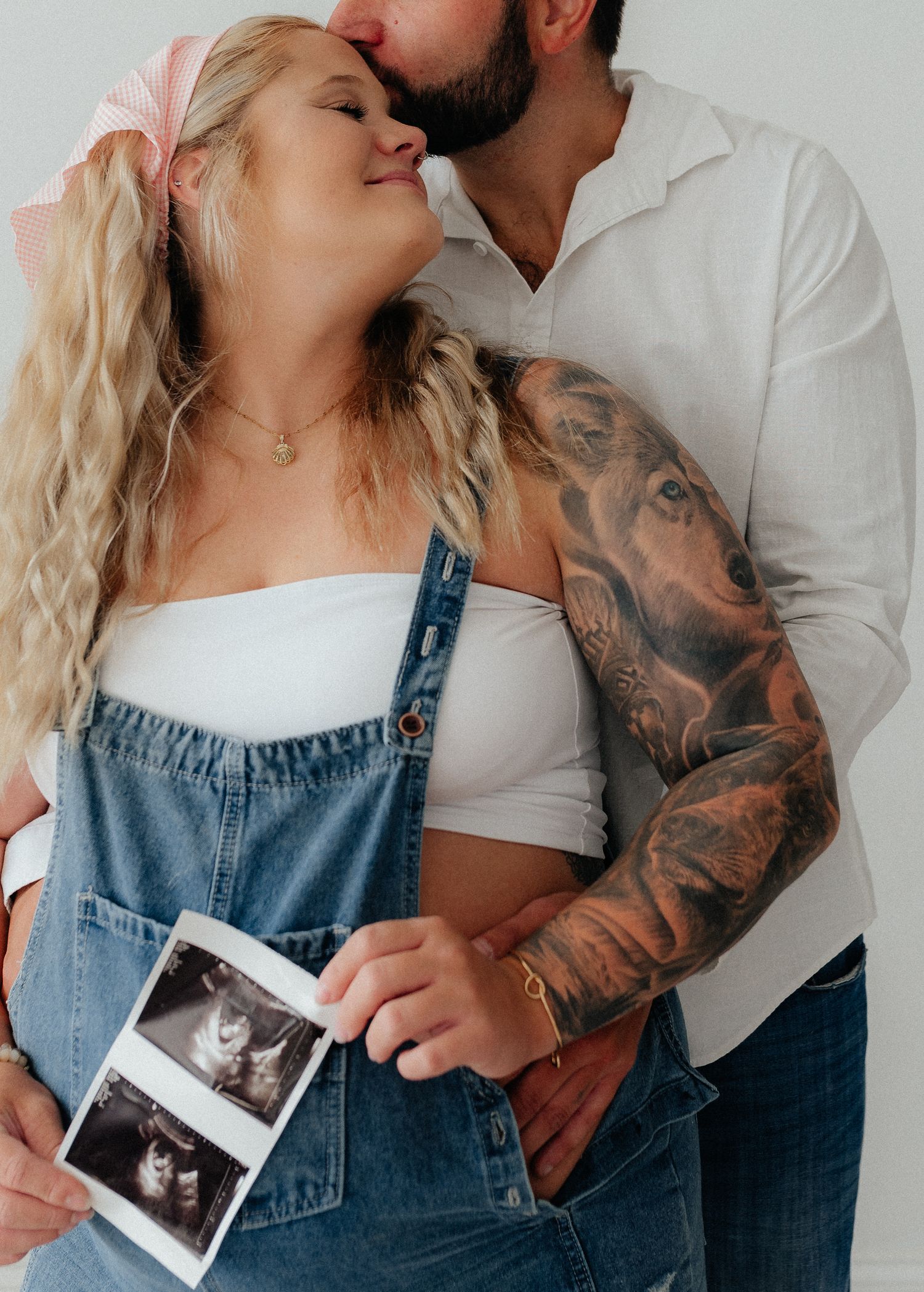 Pregnant woman in denim overalls holds ultrasound photos while partner with tattoo sleeve kisses her cheek.