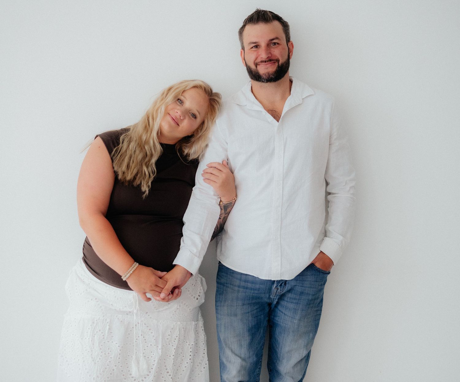 A smiling blonde woman in black top and white skirt poses with a bearded man in white shirt and jeans against a white wall.