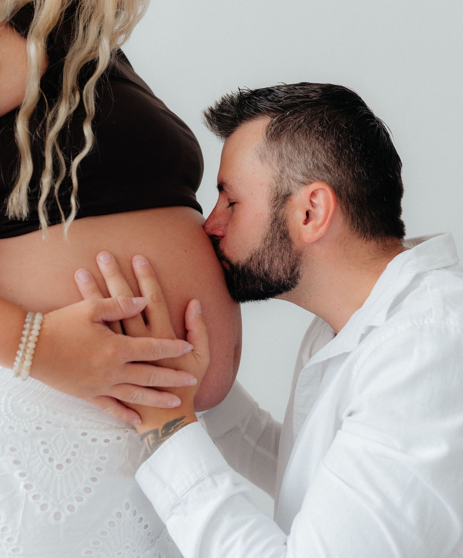 A man in a white shirt tenderly kisses a pregnant woman's bare baby bump while both gently hold it.