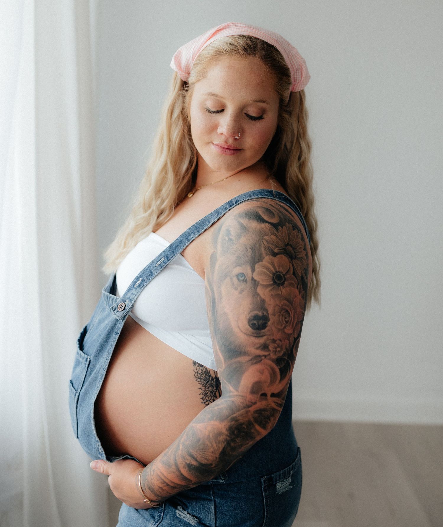 Pregnant woman with tattoo sleeve wearing denim overalls and pink headband, cradling baby bump in soft natural light.