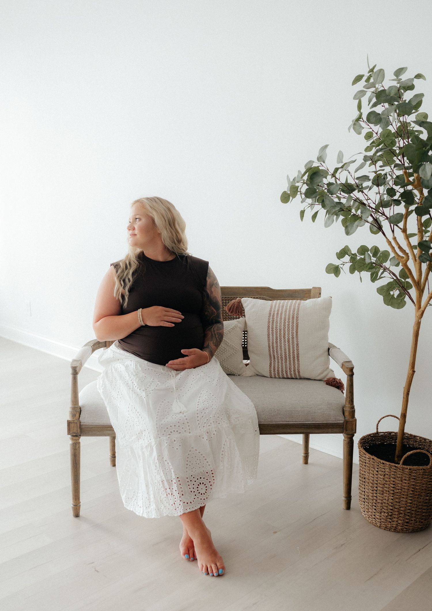 Pregnant woman in black top and white lace skirt sitting on wooden bench beside a green tree in bright studio.