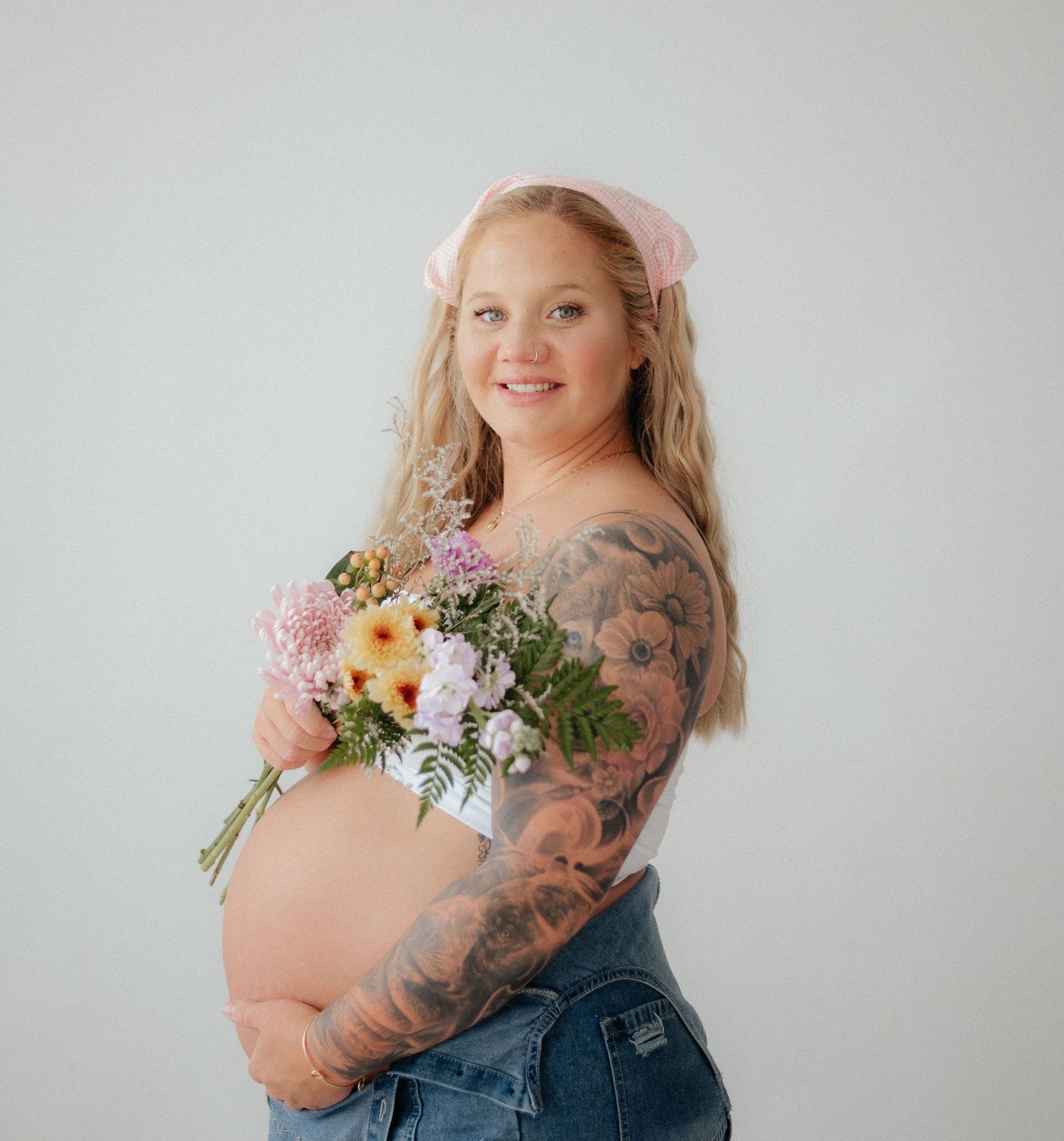 Pregnant woman with tattoos holding a pink floral bouquet, wearing jeans and a pink bow, smiling in a studio.