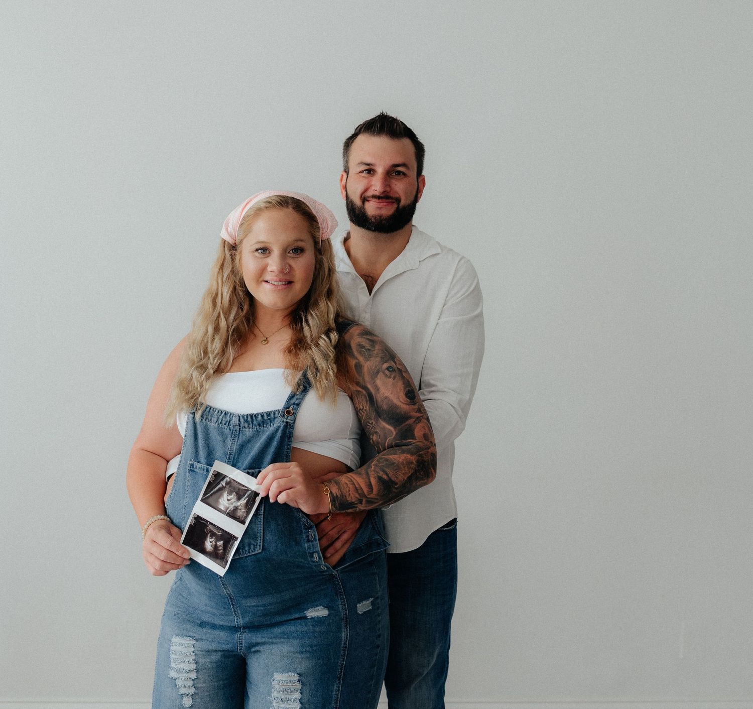 Couple holding ultrasound photos in denim overalls and white shirt, announcing pregnancy in a studio setting.