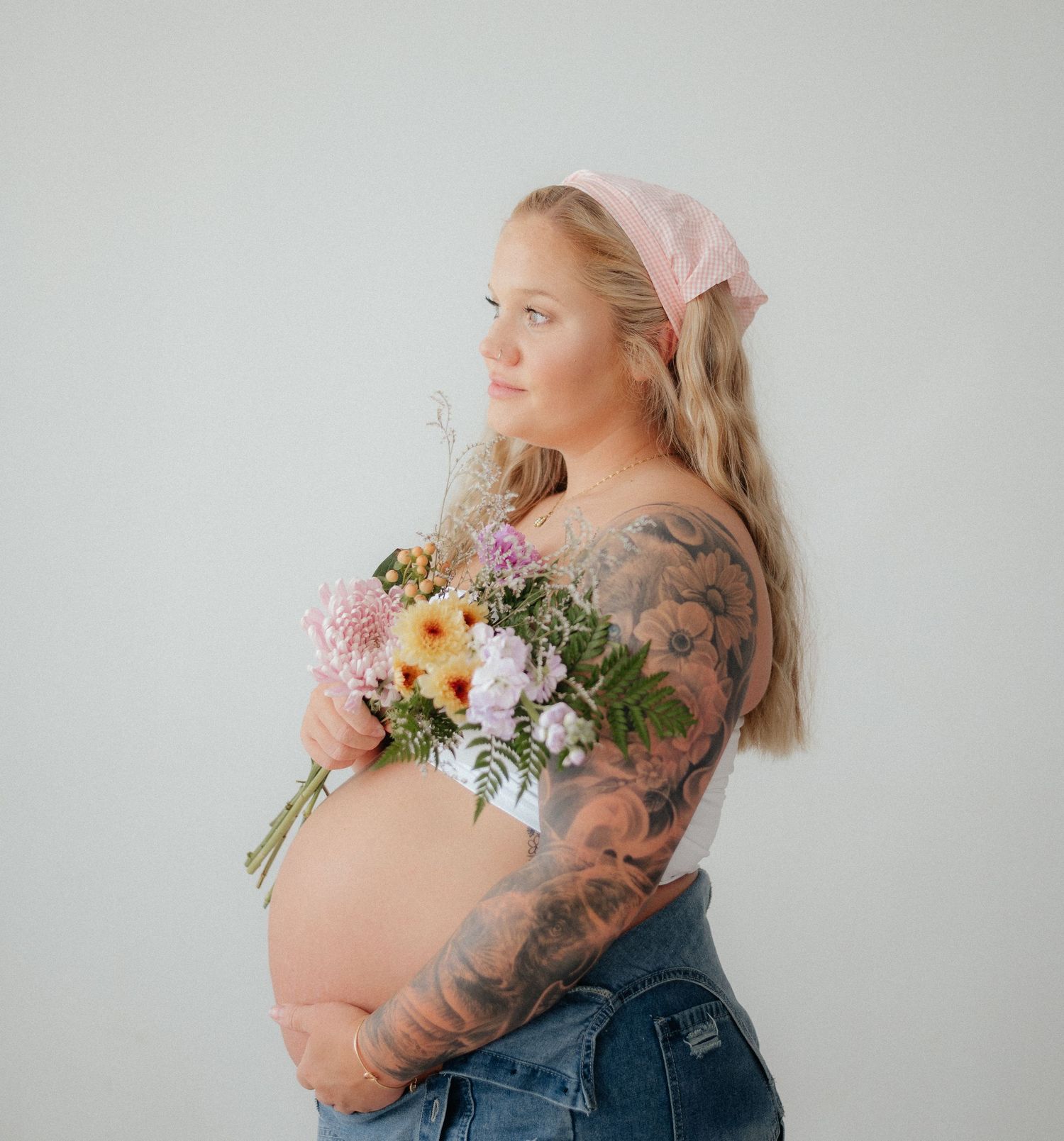 Pregnant woman with tattoos holding a bouquet of pink flowers, wearing jeans and a pink bandana, posing sideways.