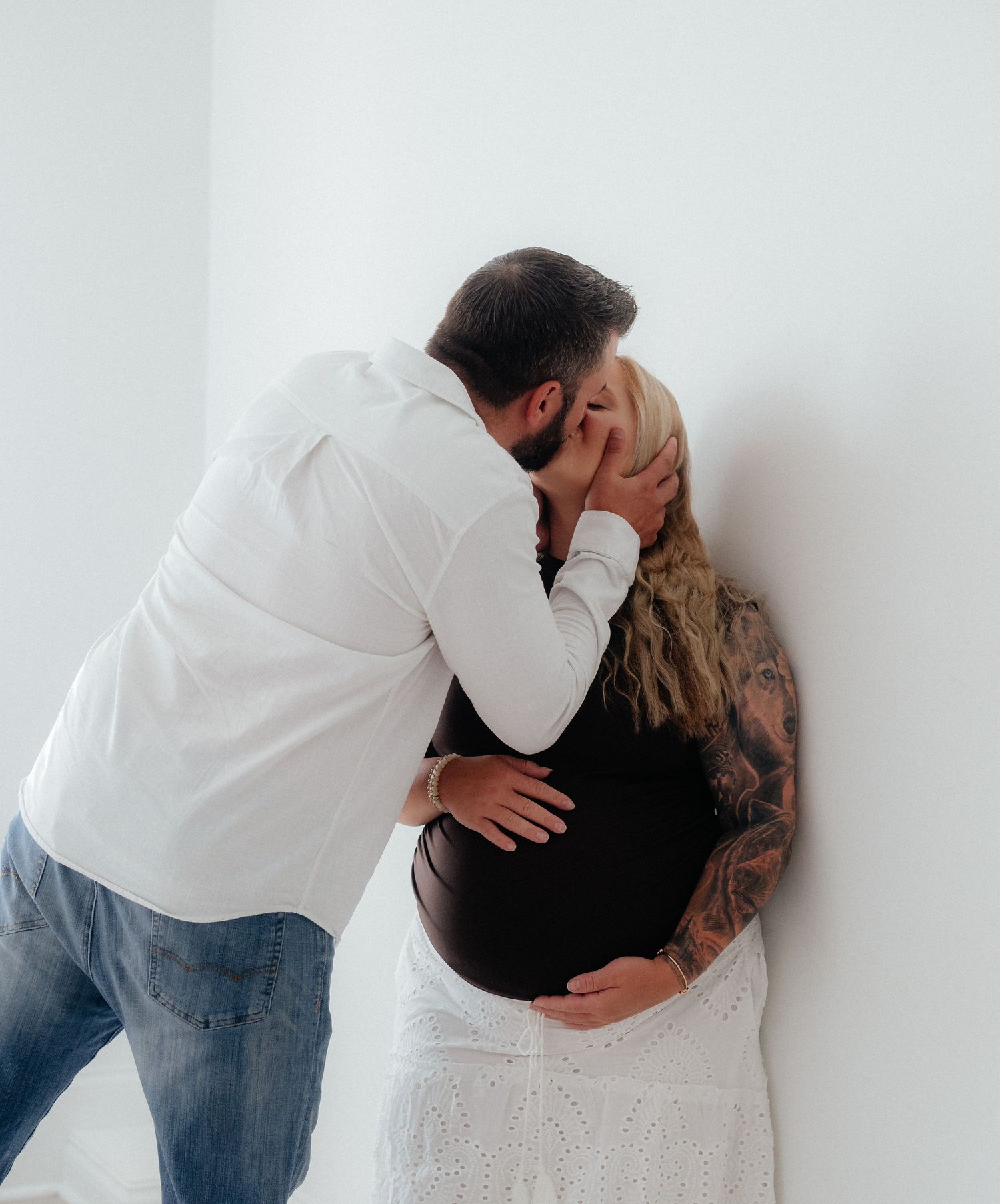 A couple shares a tender kiss against a white wall, the woman visibly pregnant with tattoos, wearing a white skirt.