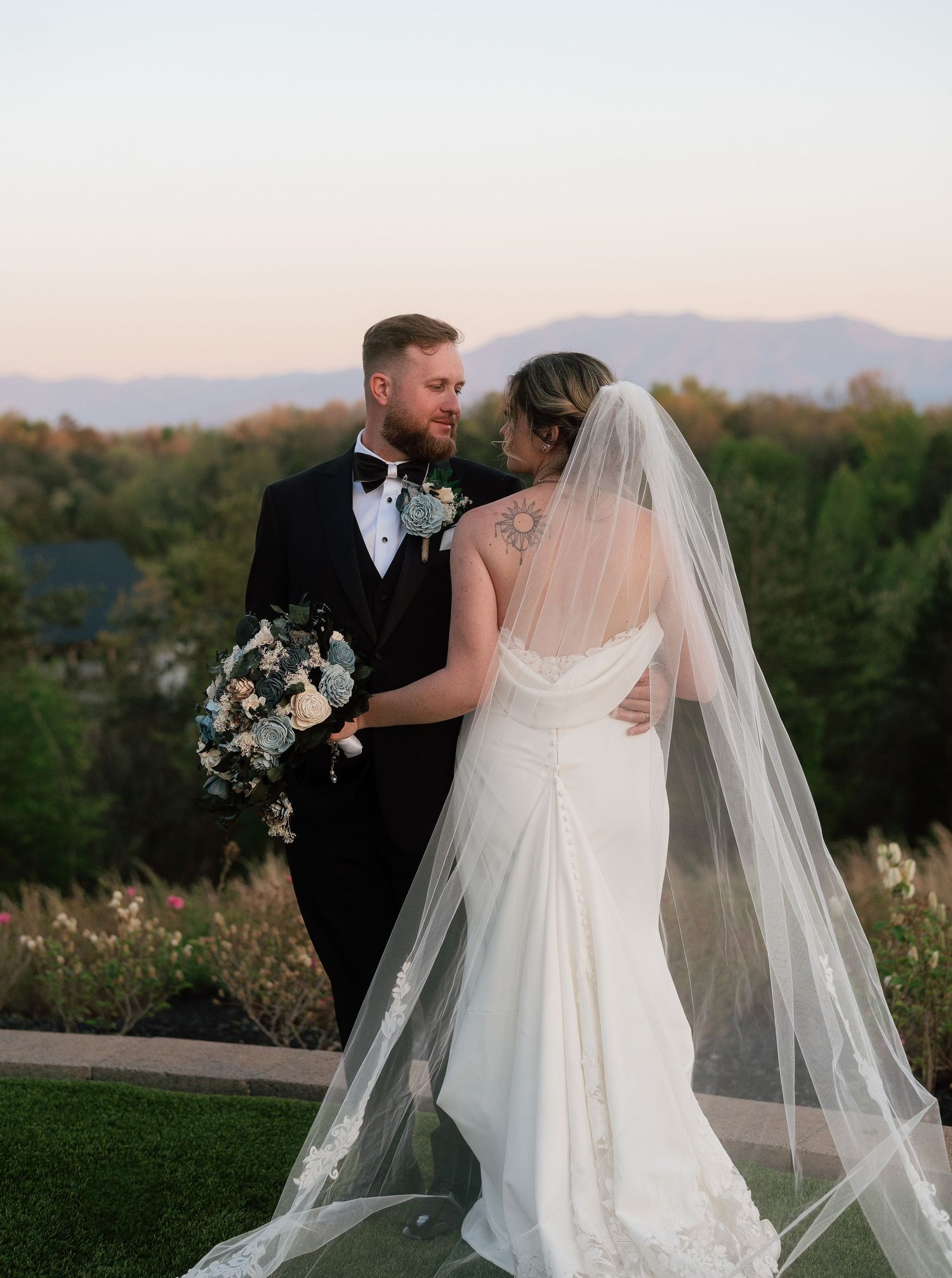 Bride and groom share a romantic moment outdoors at dusk, with mountains and gardens in the background.