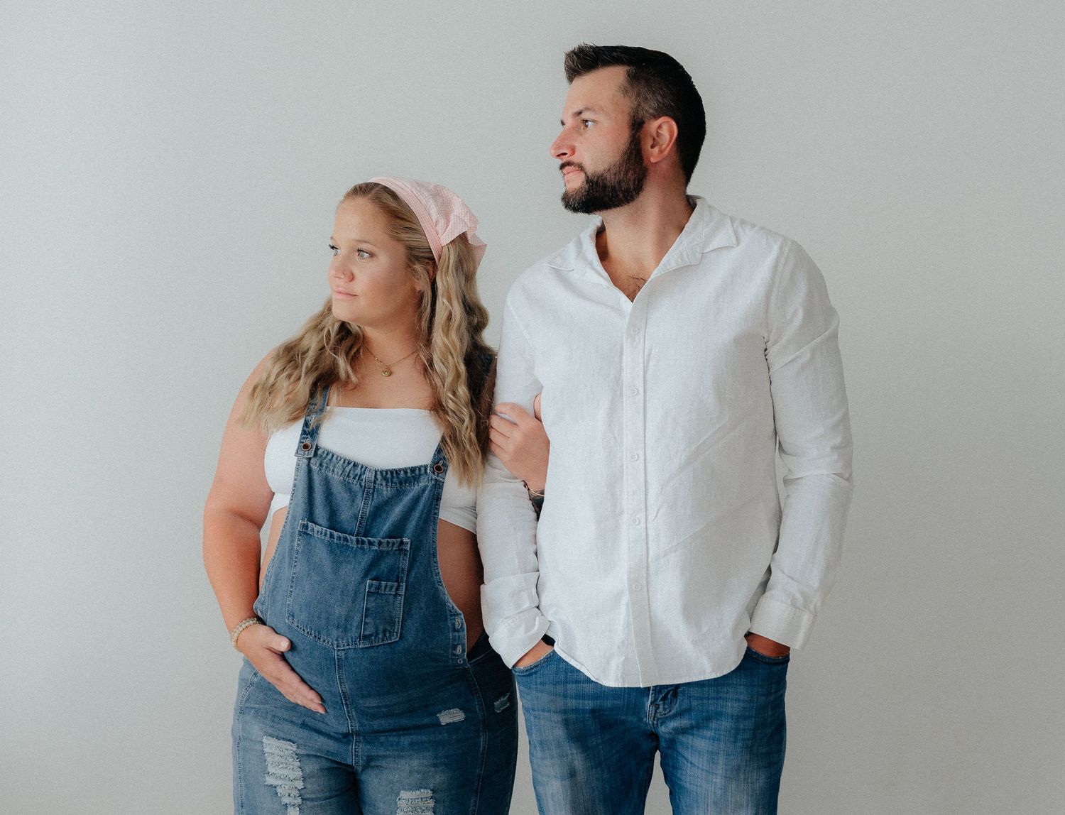 A woman in denim overalls and a man in a white shirt stand together against a light gray background.
