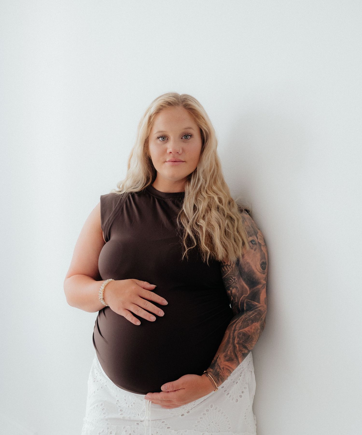 Pregnant woman with blonde hair and tattoos wearing a brown top and white skirt, cradling her baby bump against a white wall.