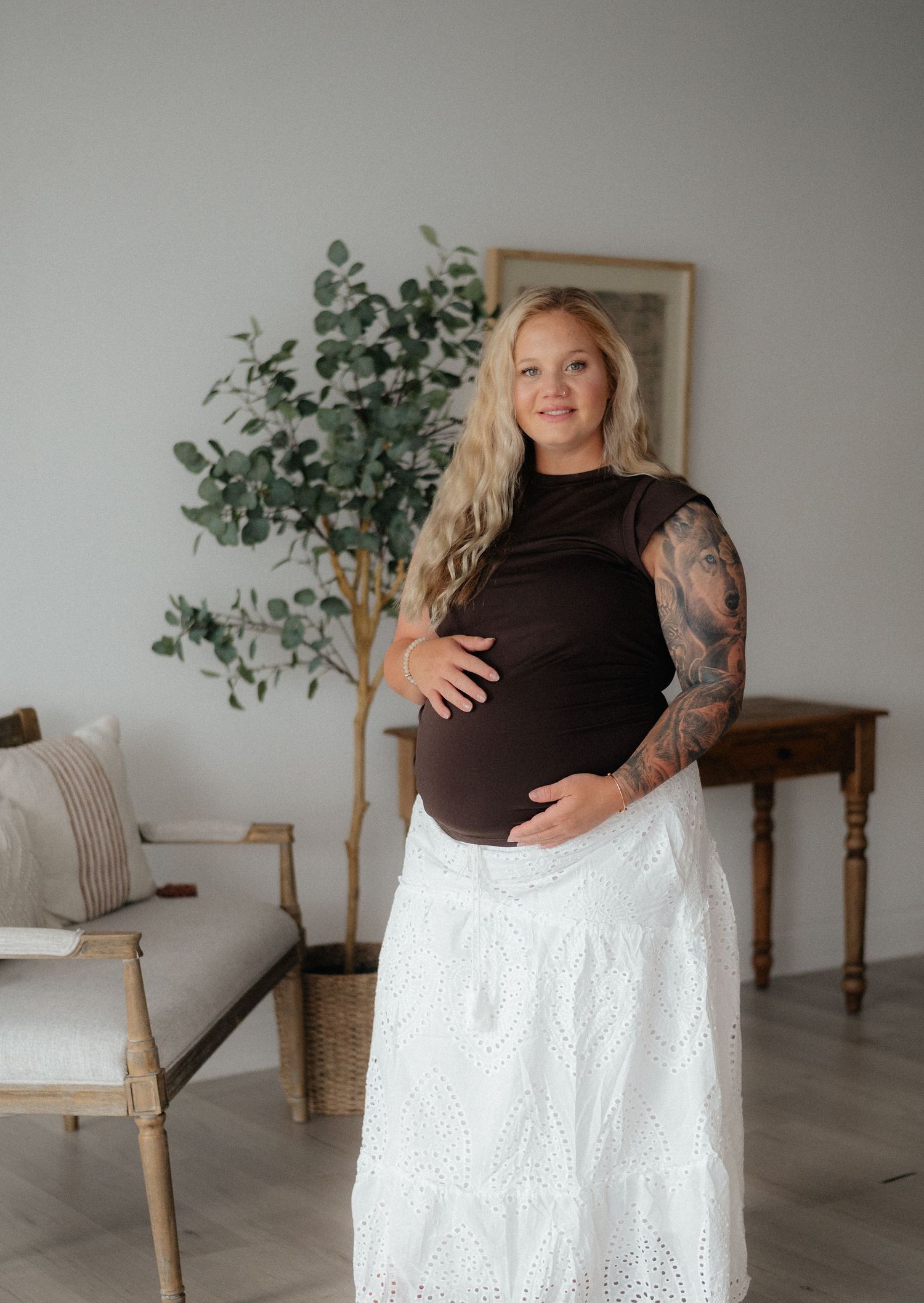 Pregnant woman in brown top and white lace skirt, showing baby bump, with tattoos, standing in bright studio setting.