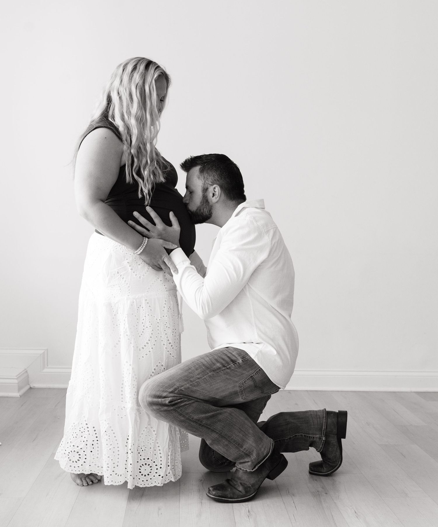 Black and white maternity photo of a man kneeling and kissing a pregnant woman's belly while she holds it gently.