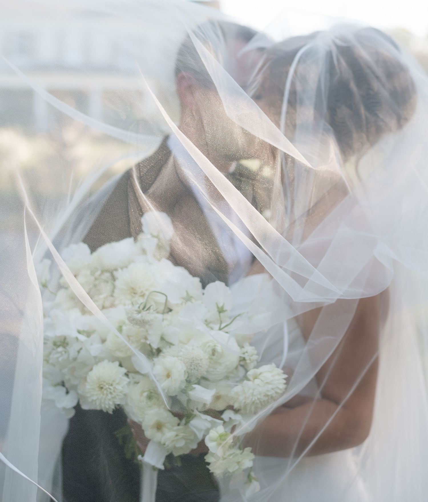 Couple shares an intimate kiss beneath a sheer bridal veil, with a white flower bouquet in soft focus.