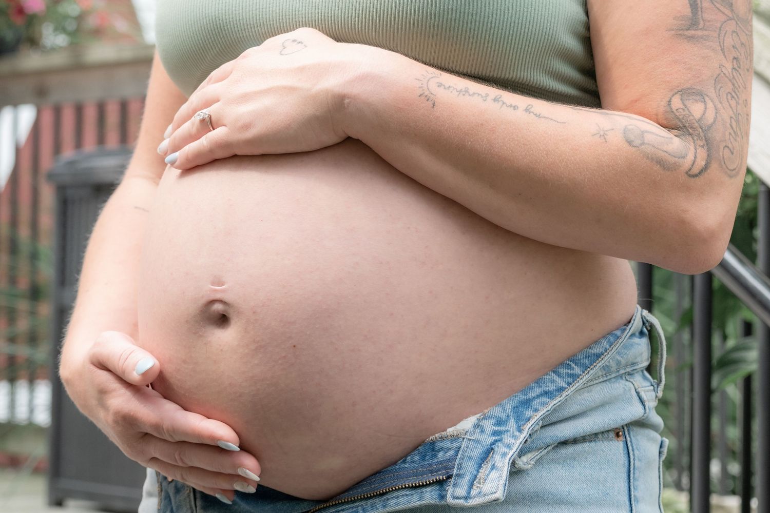 Pregnant woman cradling her bare baby bump, wearing unbuttoned jeans and a green crop top, with tattoos visible.