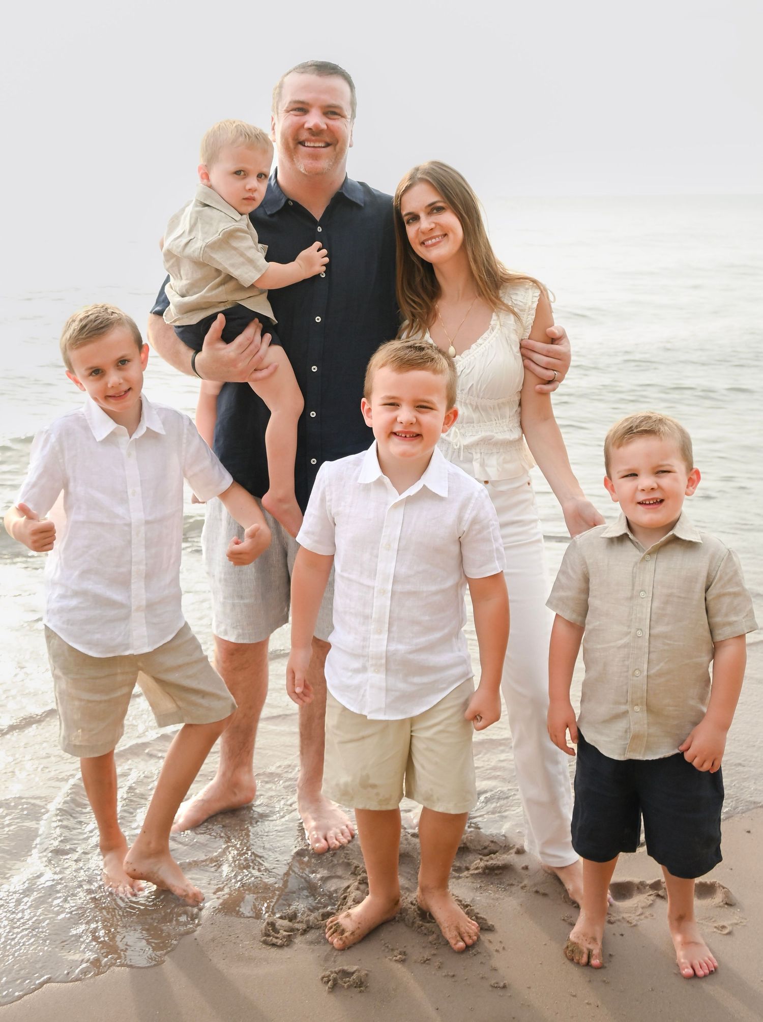 Family of six poses on a sandy beach, parents with four young boys wearing coordinating white and khaki outfits.