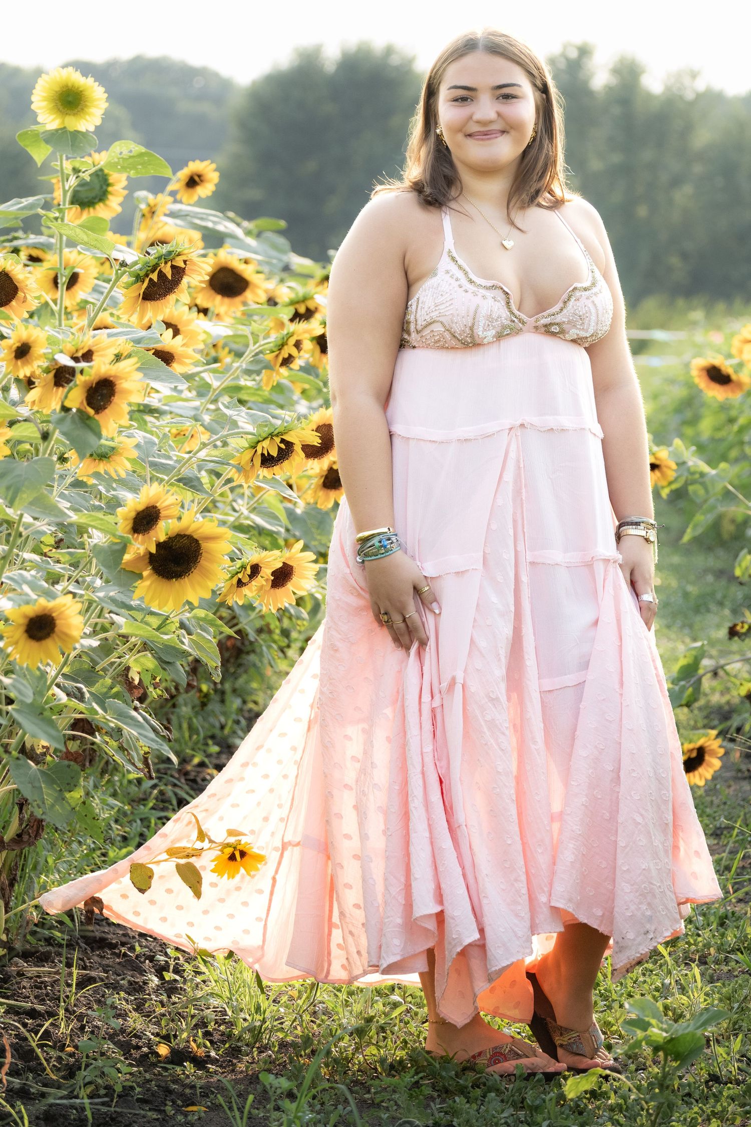 Young woman in pink maxi dress and lace bralette smiling in a sunflower field on a sunny day.