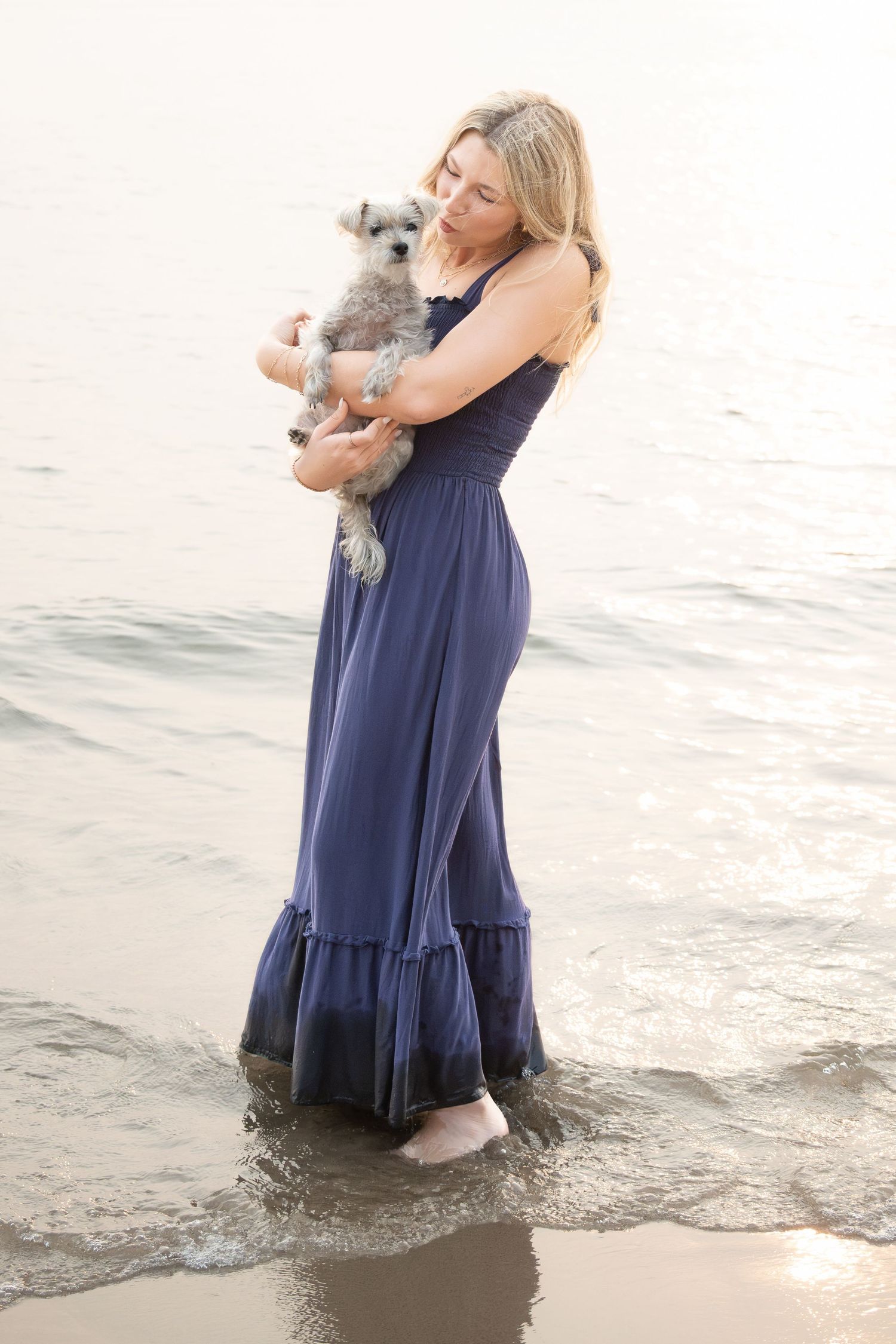 Woman in navy maxi dress holding small gray dog while standing in ocean waves at beach.