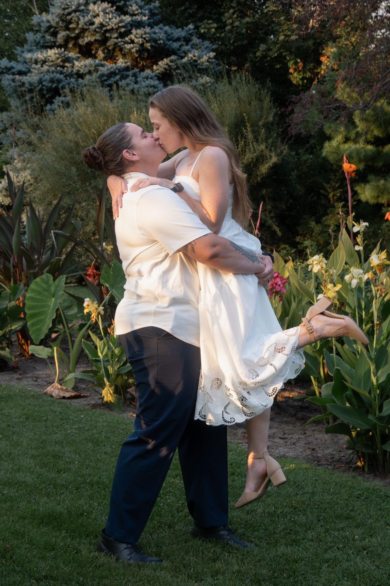 Couple sharing romantic kiss in garden, groom lifting bride in white dress among colorful flowers.