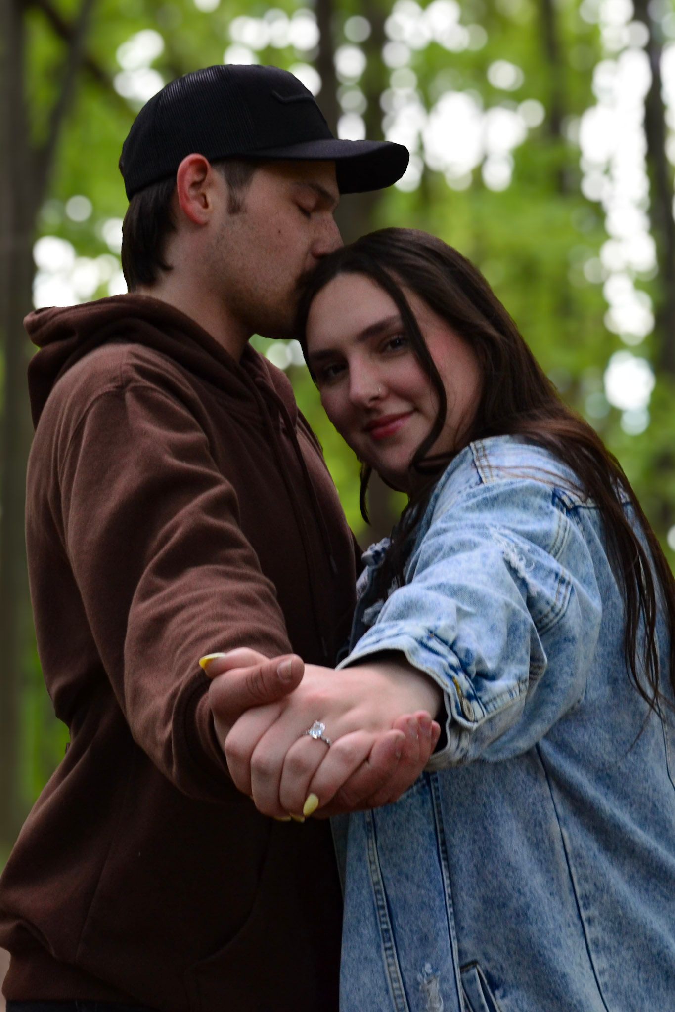 Engaged couple holding hands showing engagement ring in a green forest setting.