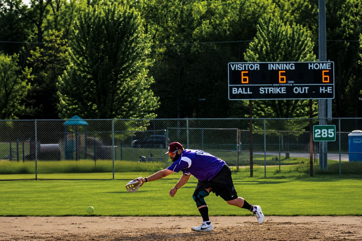 Softball player diving to field a ground ball with scoreboard showing tied game in the background.