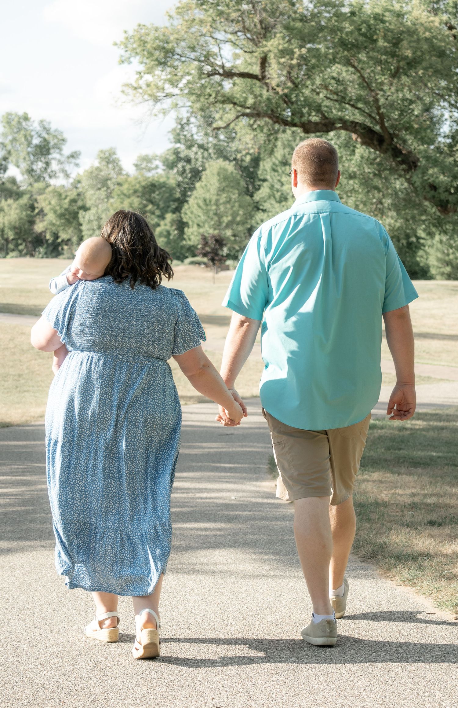 A couple walks hand-in-hand down a sunny park path, the woman in blue dress carrying a baby, man in teal shirt.