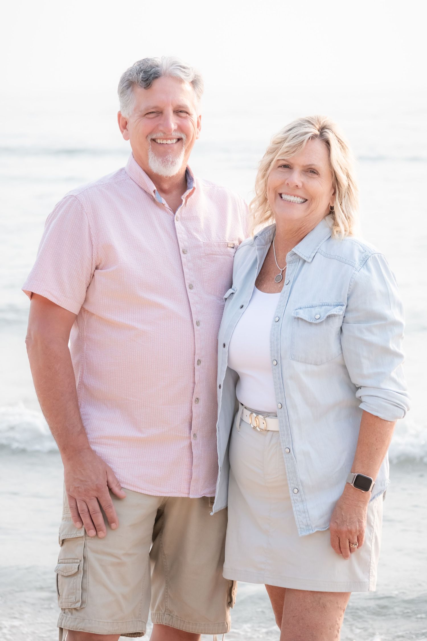 Happy middle-aged couple posing together on a beach, man in pink shirt and woman in denim jacket smiling warmly.