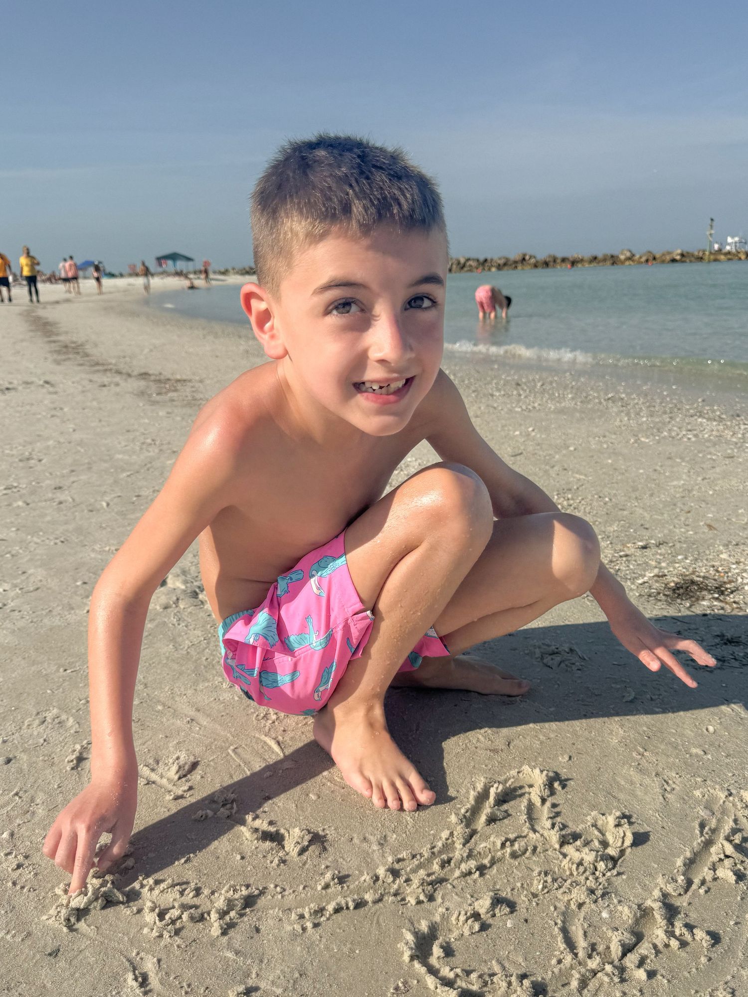 Young boy in pink swim trunks crouching and drawing in the sand at a sunny beach.