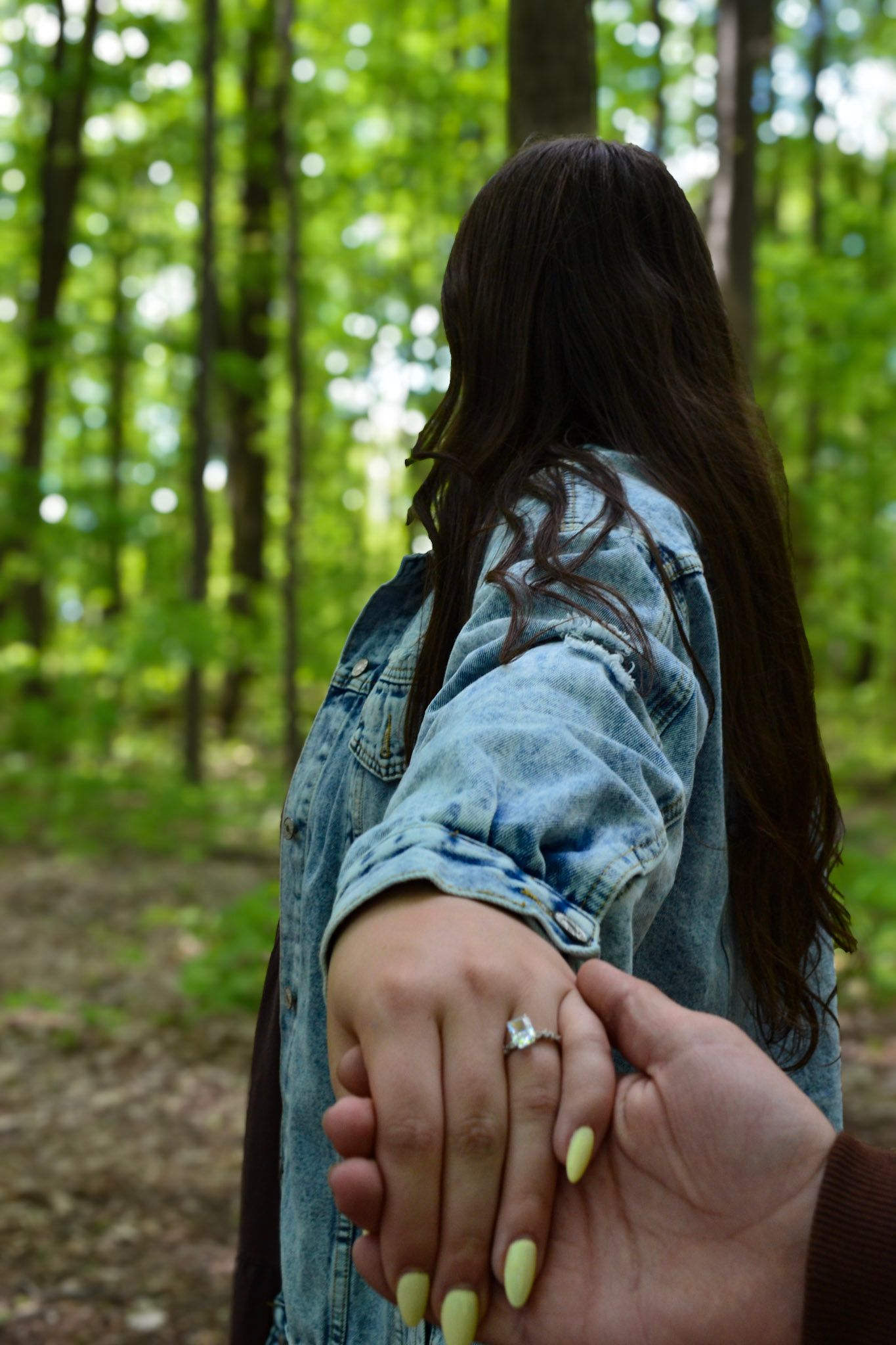 Woman in denim jacket holding hands with partner showing diamond engagement ring in forest.