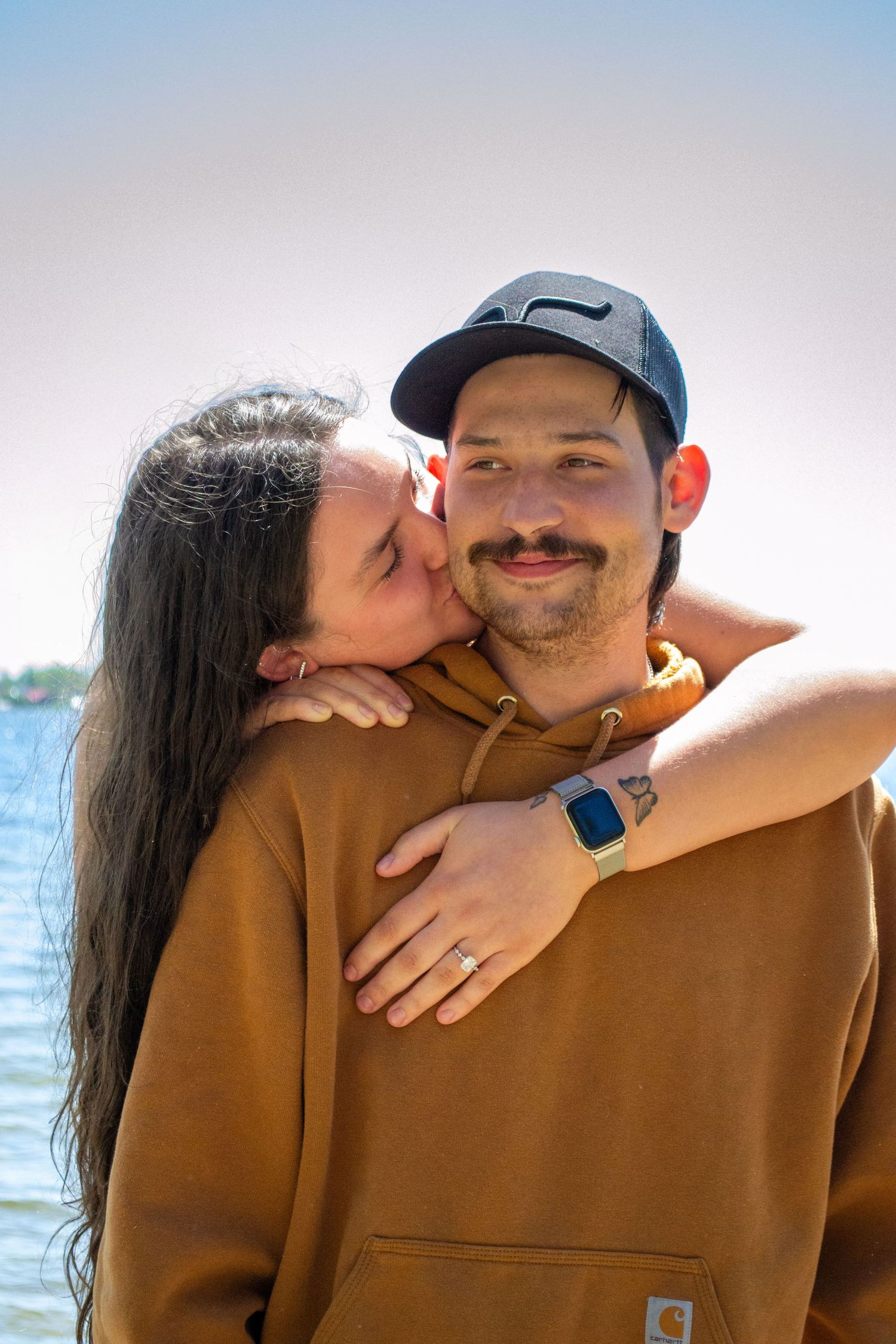 Woman kissing man on cheek near a lake, man wearing brown Carhartt hoodie and black cap.